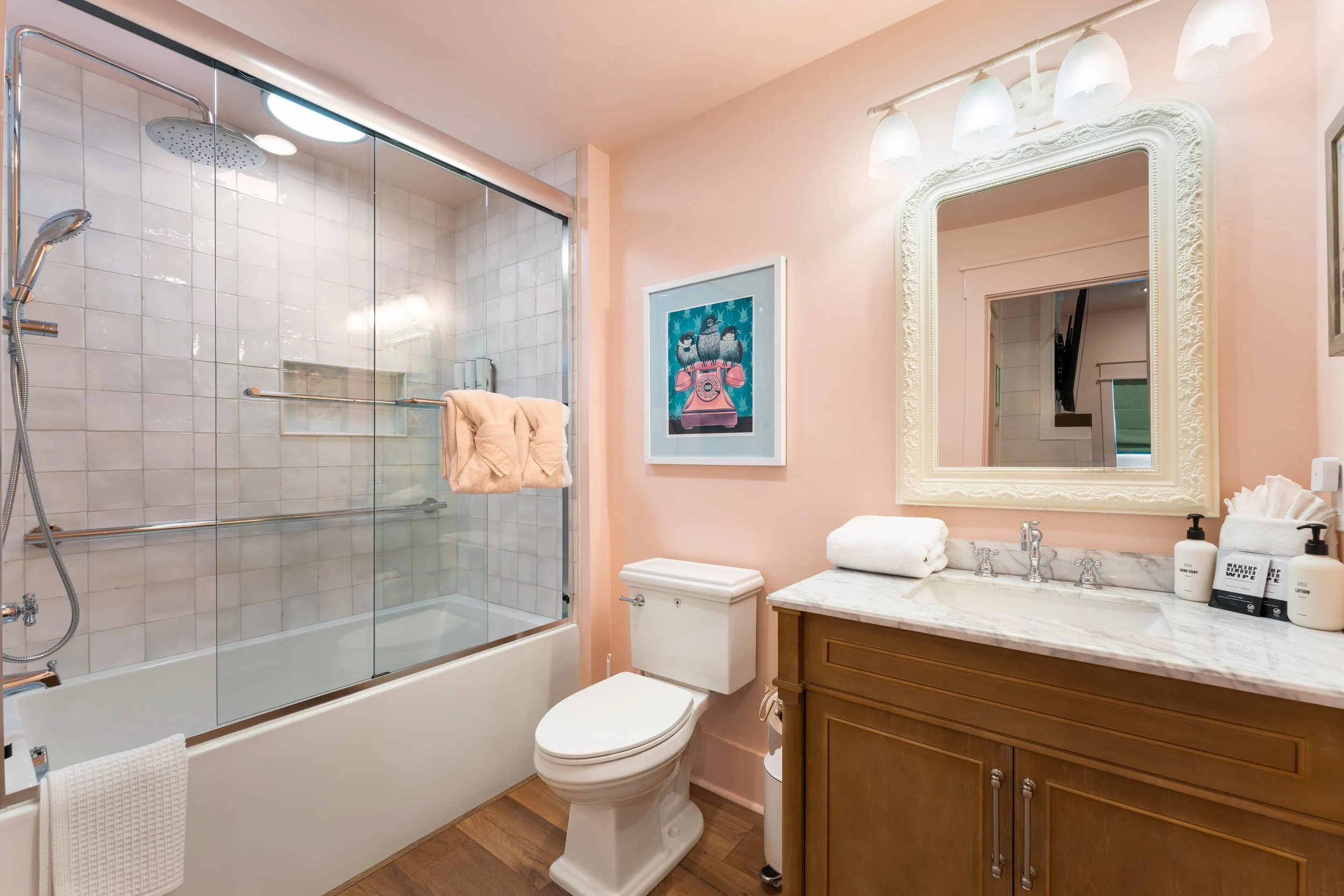 Bathroom with a glass-enclosed shower with white tiles, bathroom vanity with a marble countertop, a large mirror, framed art, and various toiletries.