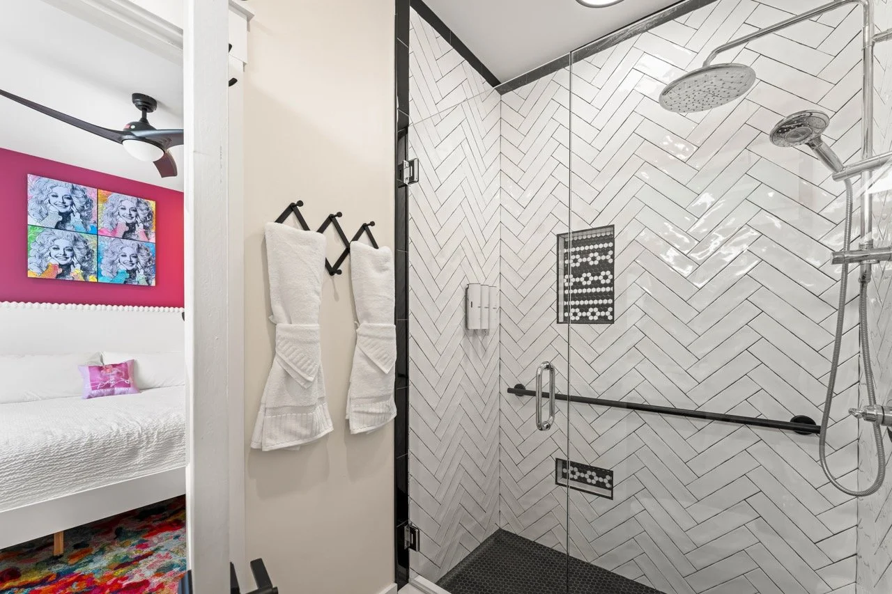 The image shows a modern bathroom with a glass shower enclosure featuring white herringbone tiles on the wall, a black grab bar, and two overhead shower fixtures. Adjacent to the shower is a wall with two white towels hanging on black hooks. In the b