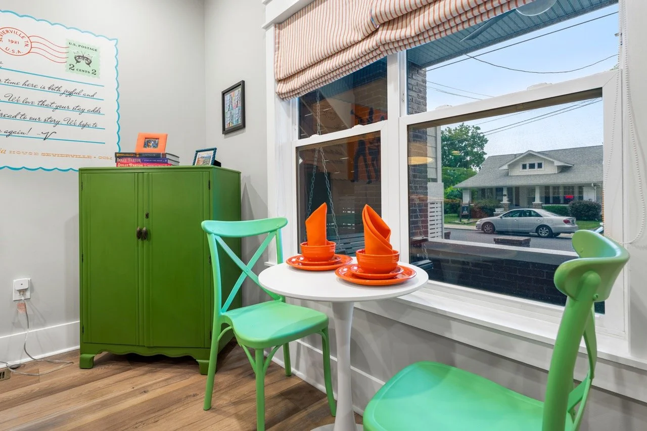 A cozy dining nook with a small white round table set with bright orange plates and folded orange napkins. Two green chairs are placed around the table. A large window with a view of a suburban street, house, and parked cars. A green cabinet with boo