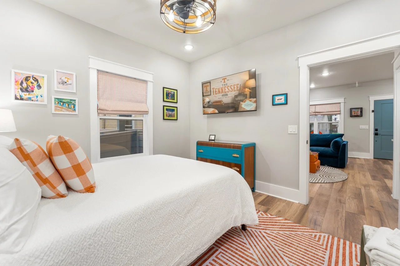 A bedroom with a white bed and orange plaid pillows, a small blue dresser, and wall art including a framed photo and a mounted TV, with a view into a living room with blue and orange furniture.