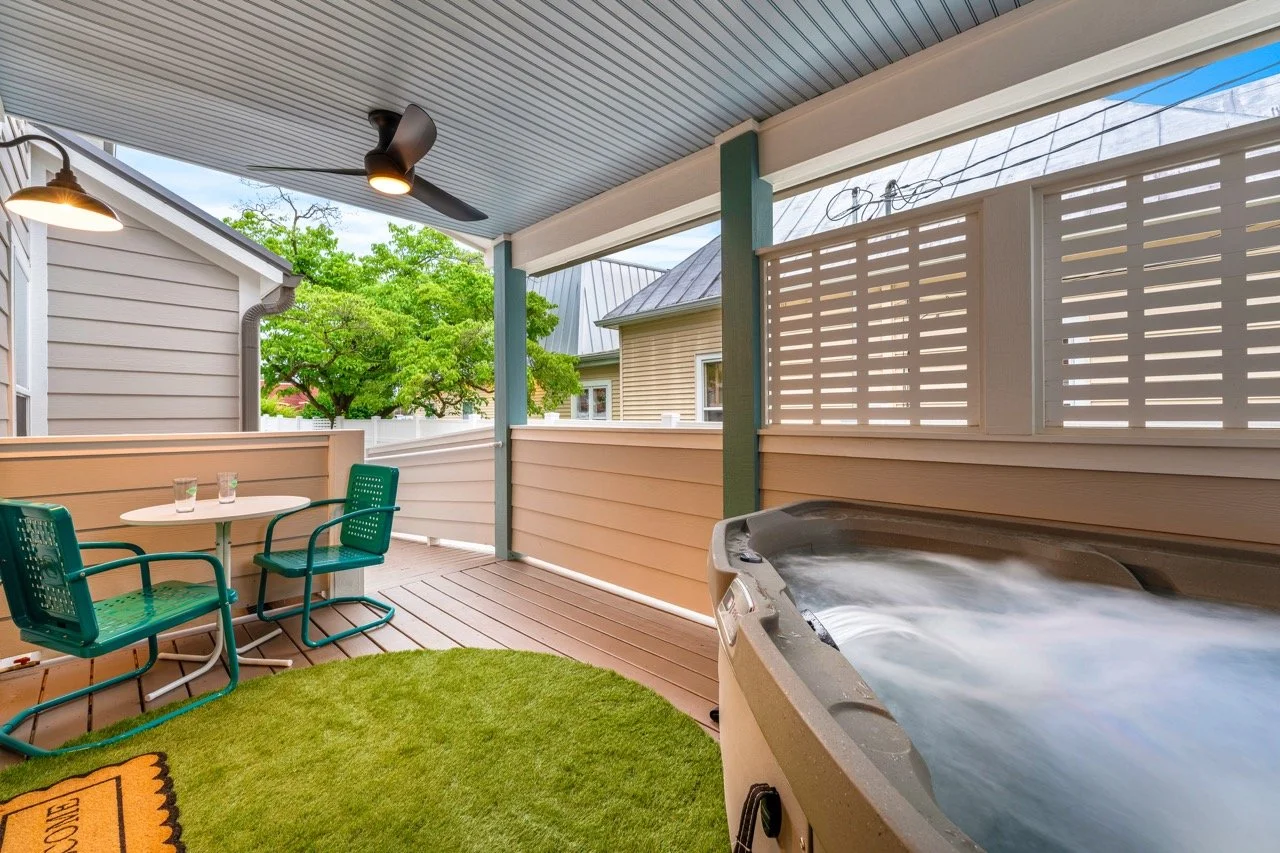 Covered backyard porch with a hot tub, a small table, and two green chairs, surrounded by a white fence and greenery.