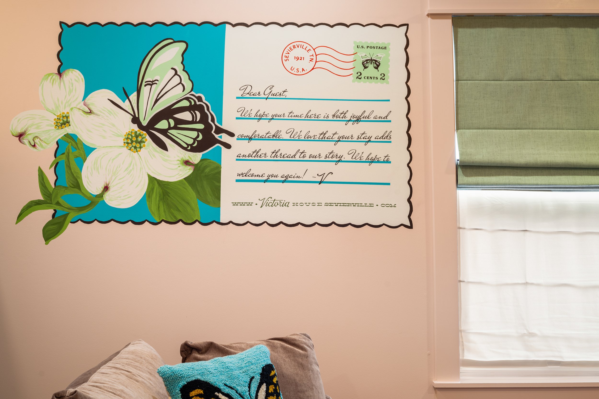 A decorative wall with a large butterfly and flower illustration next to a handwritten-style note addressed to a guest, stamped with vintage postage.