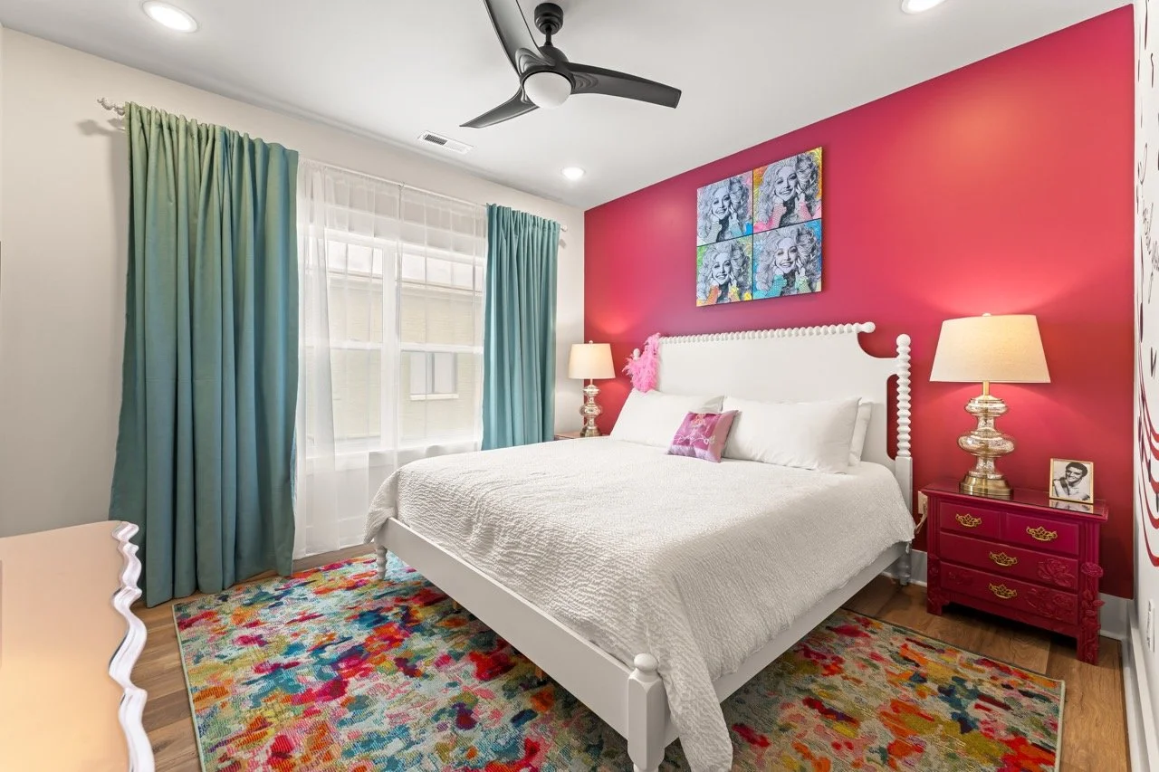 A bedroom with a white bed against a pink accent wall, colorful artwork above, teal curtains, a colorful area rug, gold and pink lamps, and a framed photo on a pink nightstand.