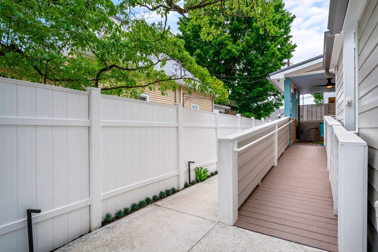 View of a backyard with a concrete patio, white fence, and a wooden porch with a ramp attached. Green trees are visible over the fence, and the porch has a ceiling light.