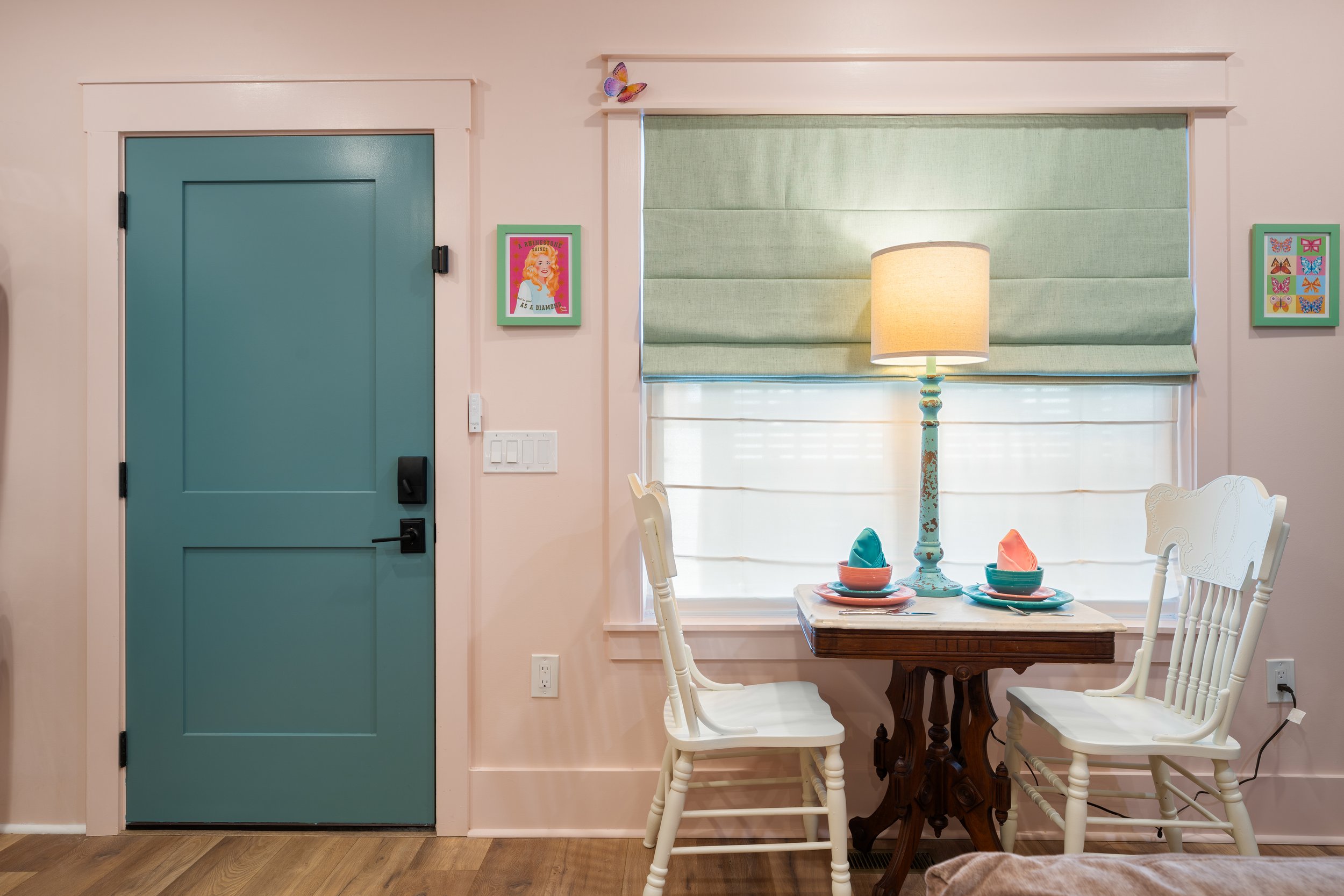 A wall with a teal door on the left and a window with a green shade on the right. There is a table with four chairs, set with teal napkins and dishes, and a vintage table lamp with a beige shade. There are framed pictures, including a small one of a 