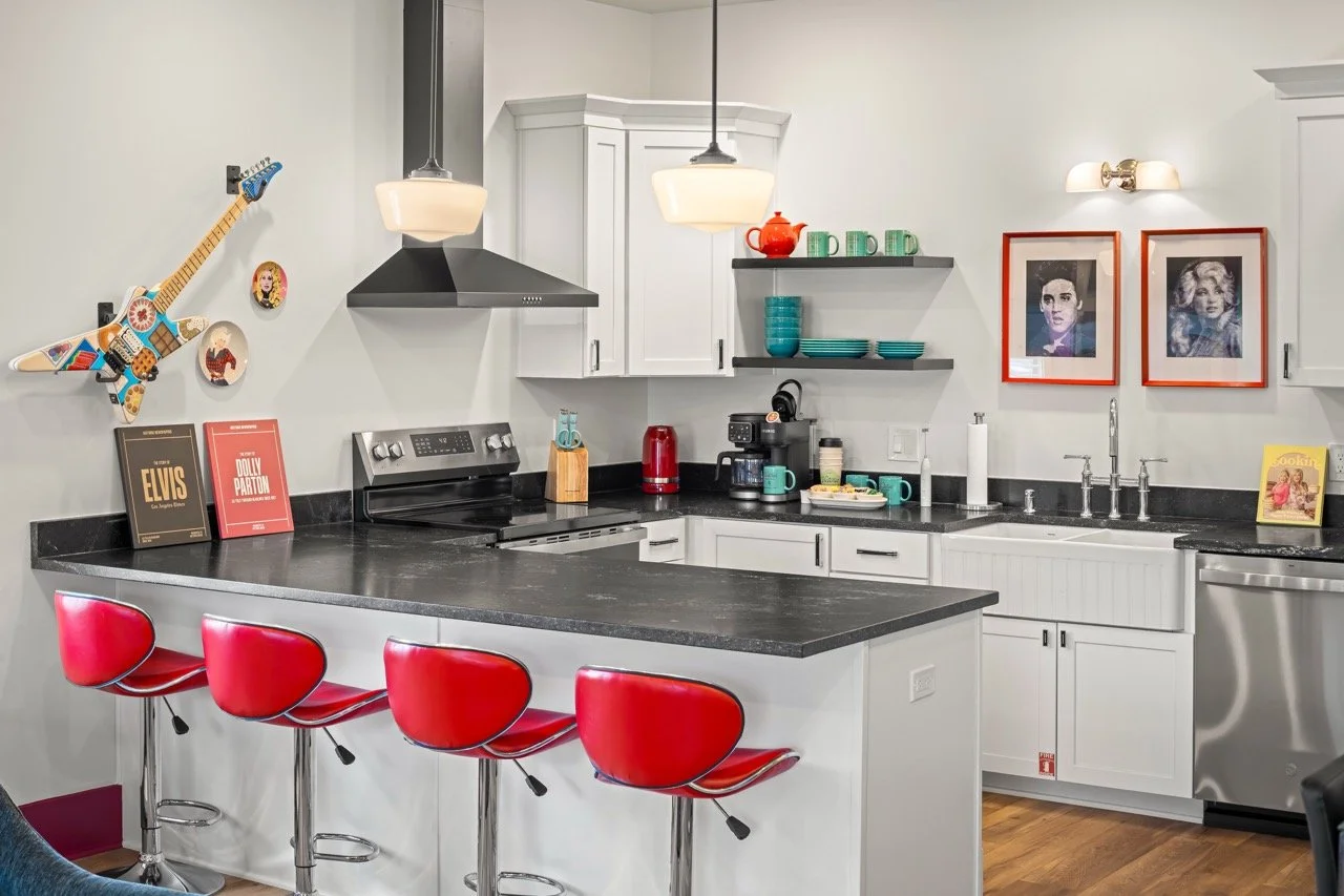 Kitchen with white cabinets, black countertops, a black range hood, and red barstools. Decor includes a guitar wall hanging, framed portraits, and various teal and red dishware.