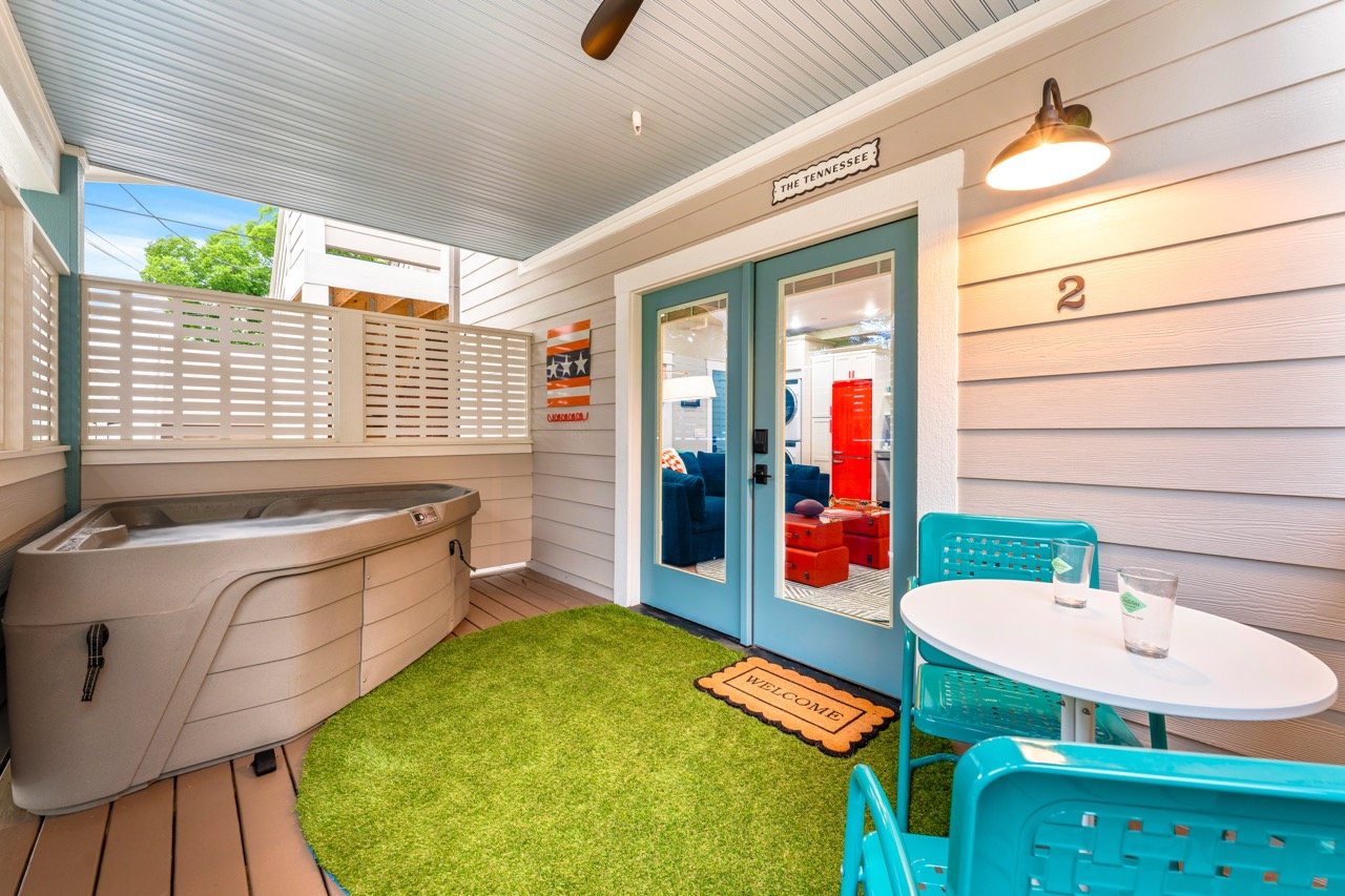 Covered outdoor porch featuring a hot tub, a small round table with two blue chairs, two glasses of water on the table, a welcoming doormat, and a door leading into a living room. The porch has white siding, a ceiling fan, and a sign that reads 'The 