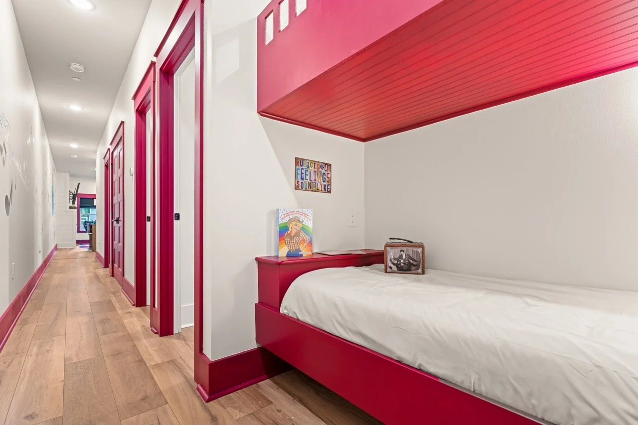 Child's bedroom with a built-in red bed under a red shelf, white walls, colorful wall art, and a wooden floor.