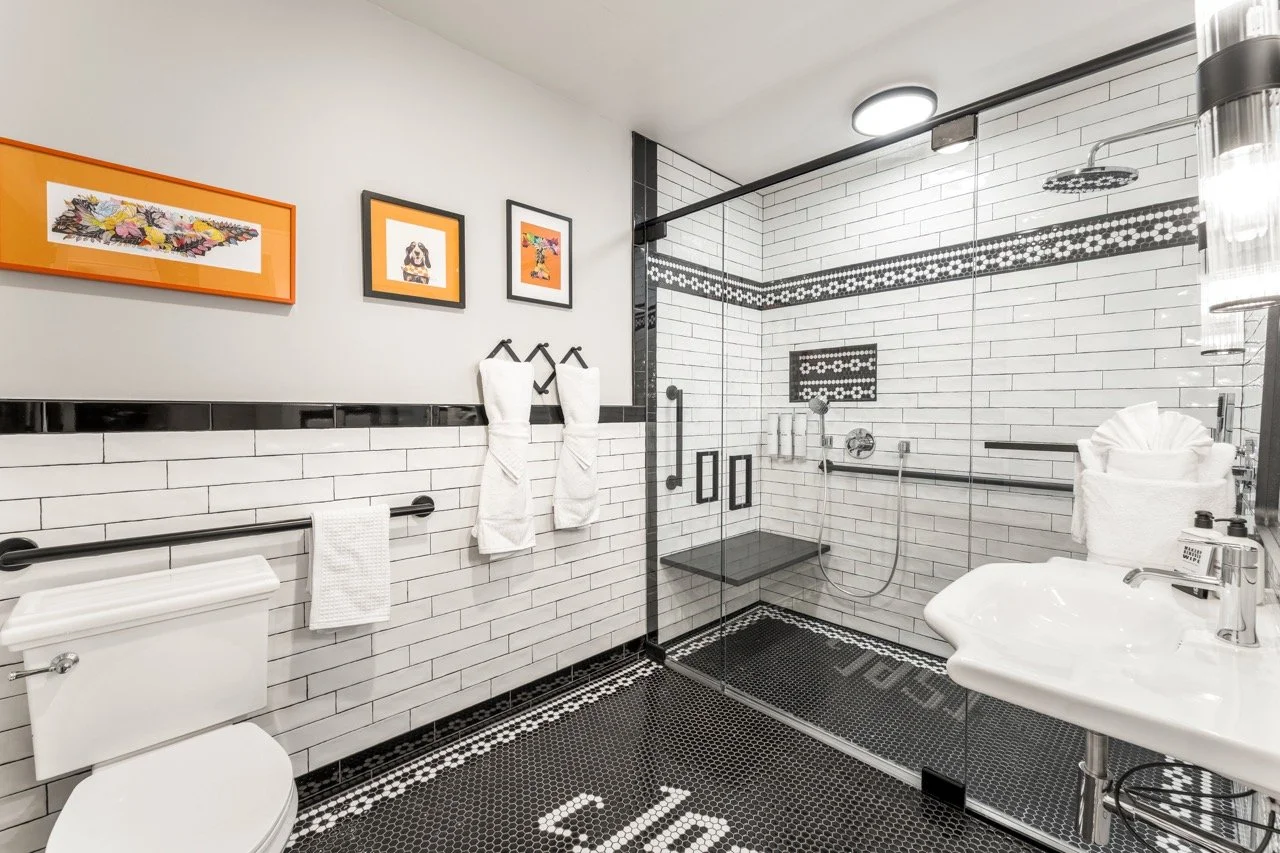 Modern bathroom with white subway tiles, black accents, and artwork, featuring a walk-in shower with glass door, black non-slip floor, toilet, and a small sink with toiletries.