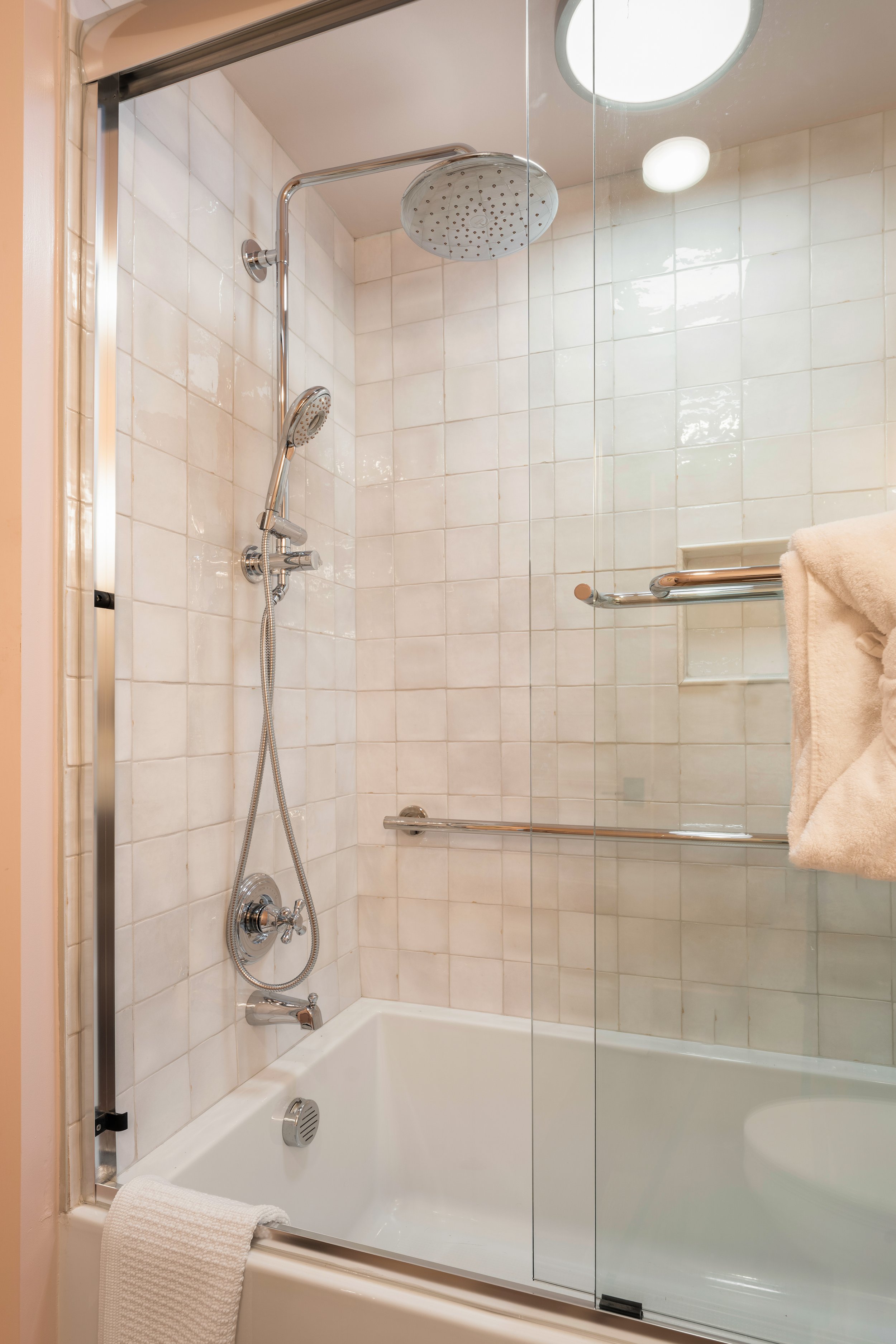 A bathroom shower with white tiled walls, a large overhead showerhead, a handheld showerhead, and a glass door. There is a towel hanging over the shower door.