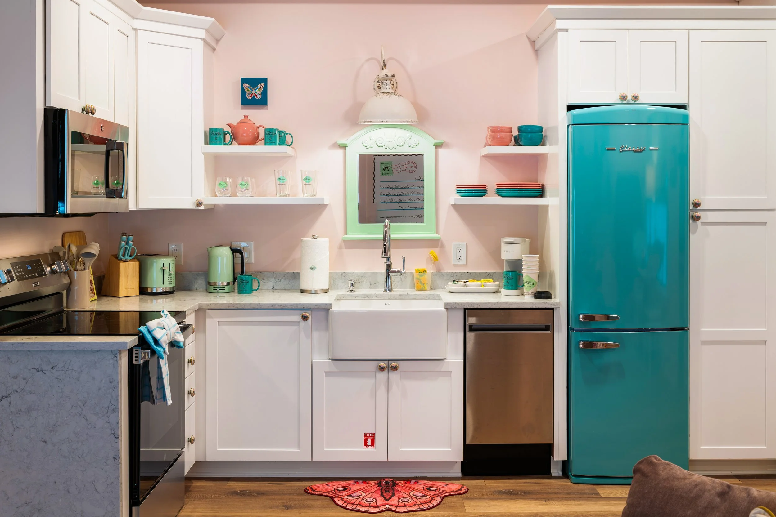 A colorful kitchen with white cabinets, a teal refrigerator, and a pink wall. Various kitchen items such as cups, bowls, and a paper towel roll are on the countertop. There is a pink and orange butterfly rug on the floor and a brown dog at the bottom right corner.