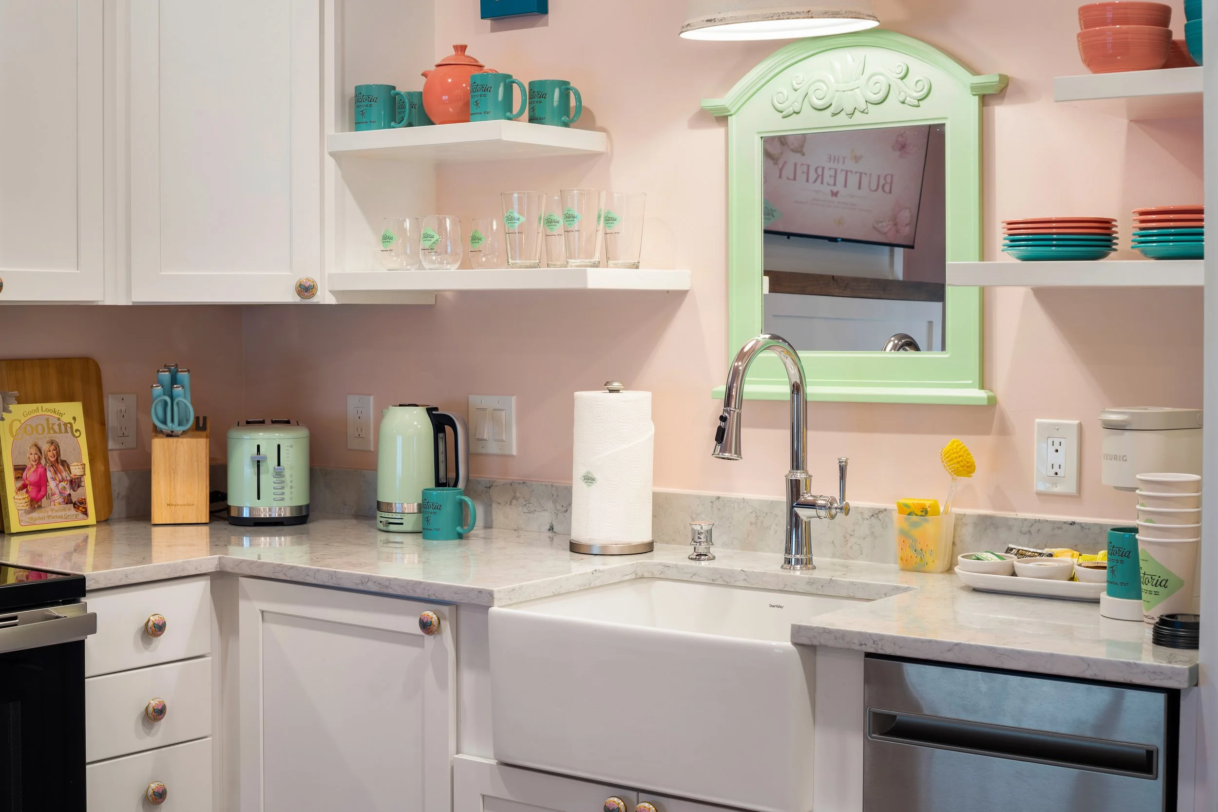 A kitchen with white cabinets, a marble countertop, and open shelves holding colorful dishes and cups. There is a sink, a paper towel holder, and small kitchen appliances like a toaster and kettle. A large green-framed mirror is mounted on the wall, 