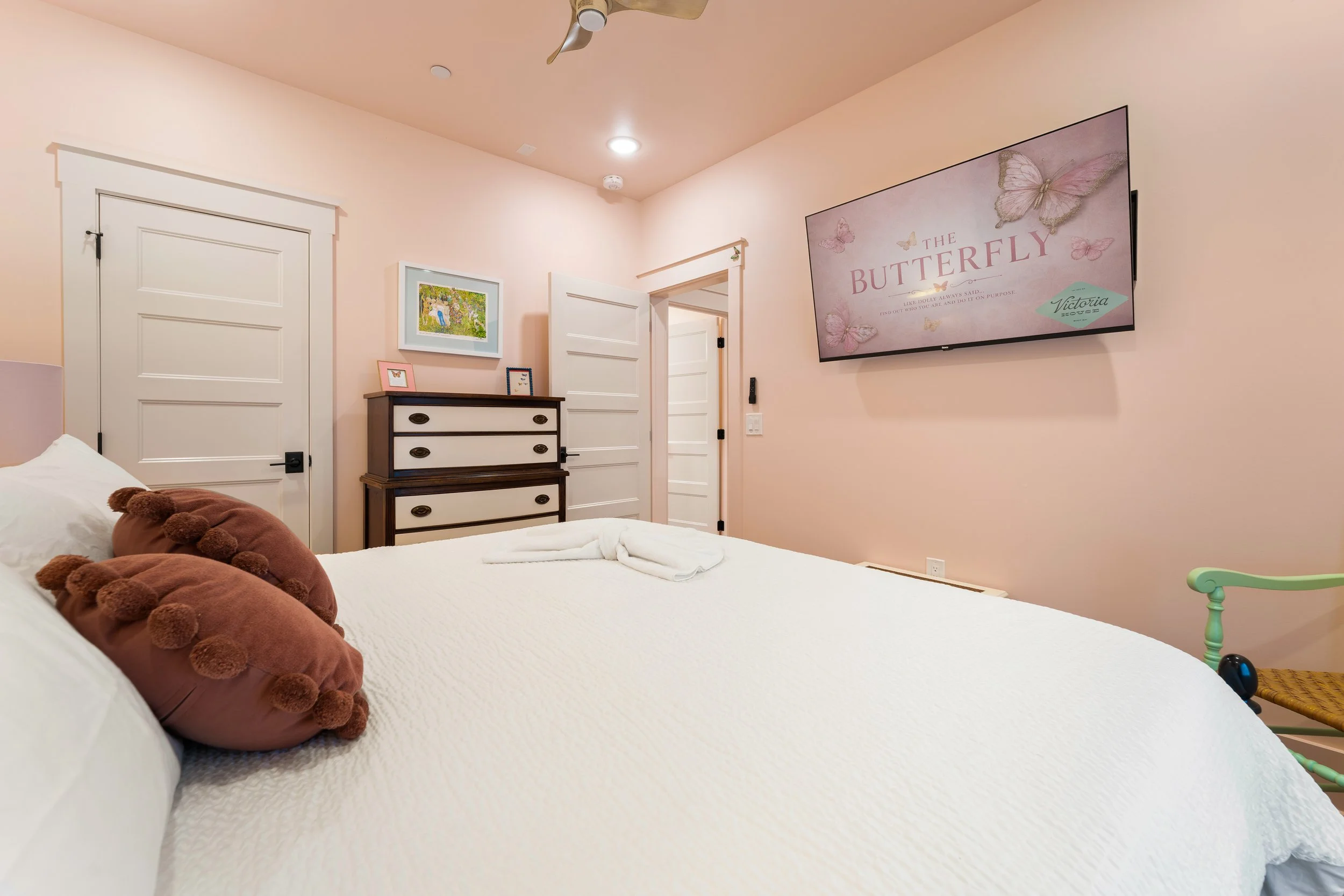 A bedroom with a bed covered in white bedding and a brown, pom-pom decorated pillow. The walls are painted light pink. There are two closed white doors, a dark wooden dresser with framed pictures on top, and a large flatscreen TV mounted on the wall 
