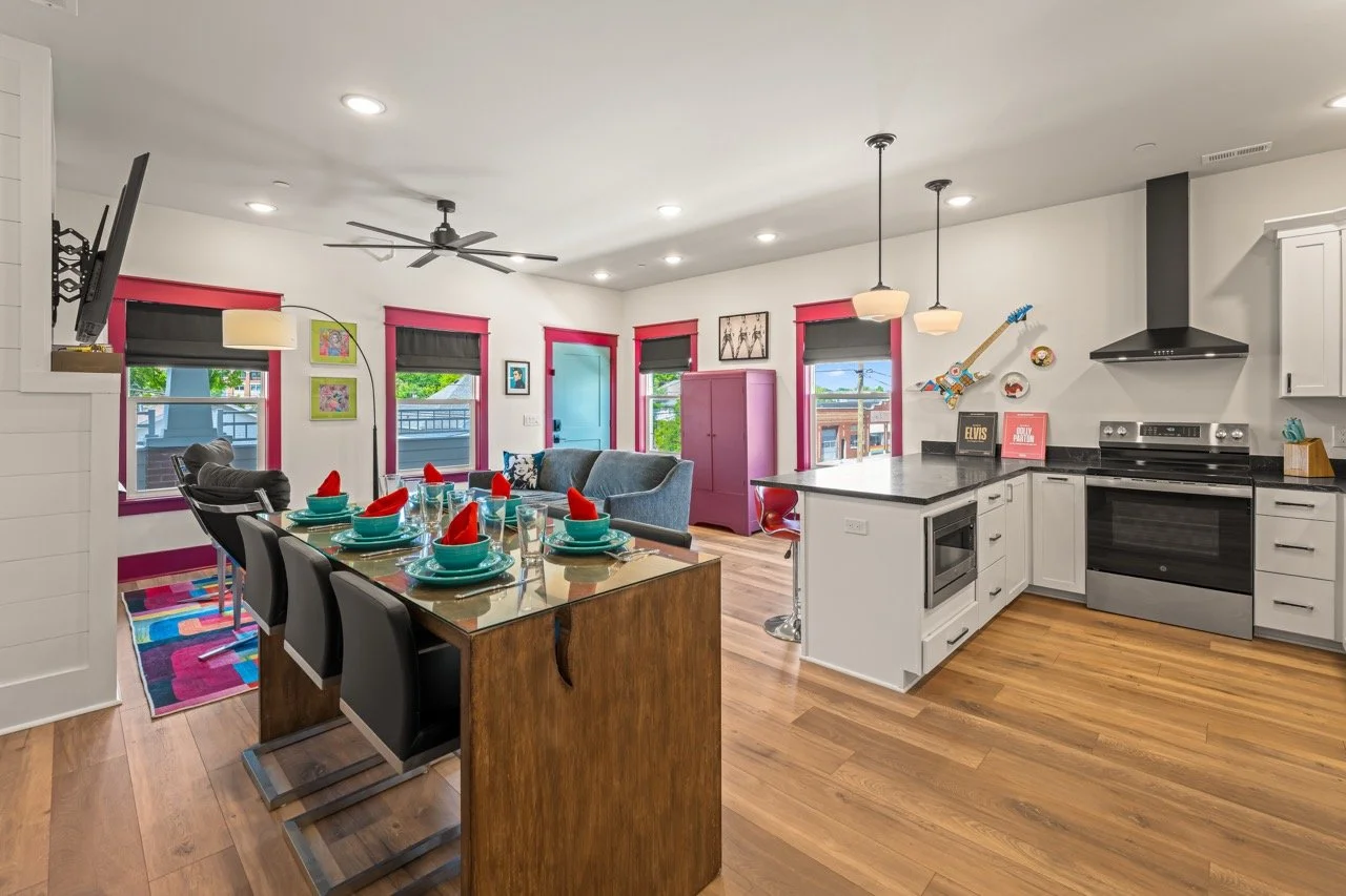 Open-concept kitchen and dining area with a wooden dining table set for a meal, black chairs, a living room with a blue couch, multiple windows with pink and black blinds, and modern appliances.