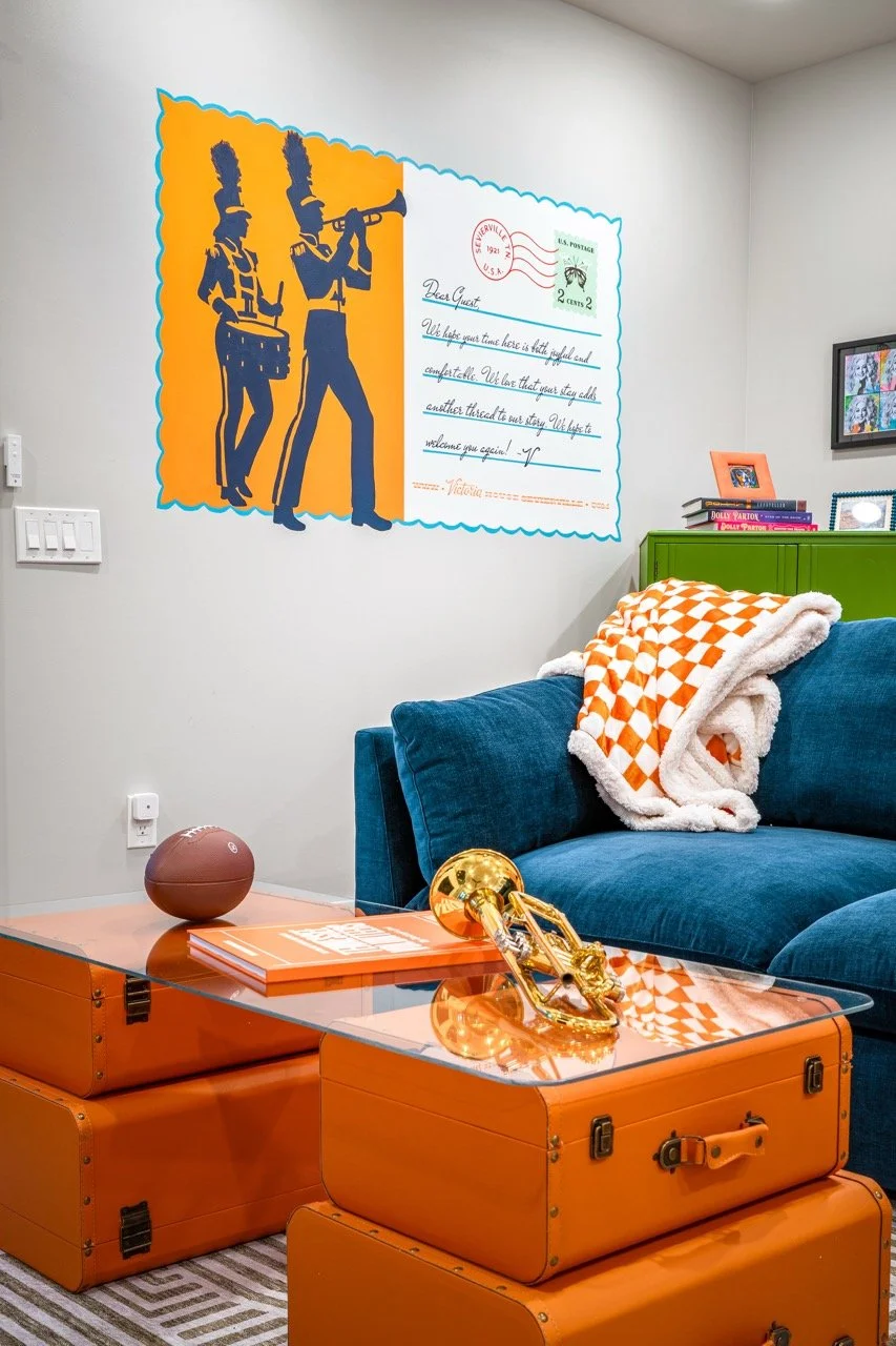 Living room with orange vintage suitcases used as a coffee table, a blue velvet sofa with a blanket, a green cabinet with books and picture frames, and a wall mural of two marching band figures, a letter with a postage stamp, and handwritten notes.