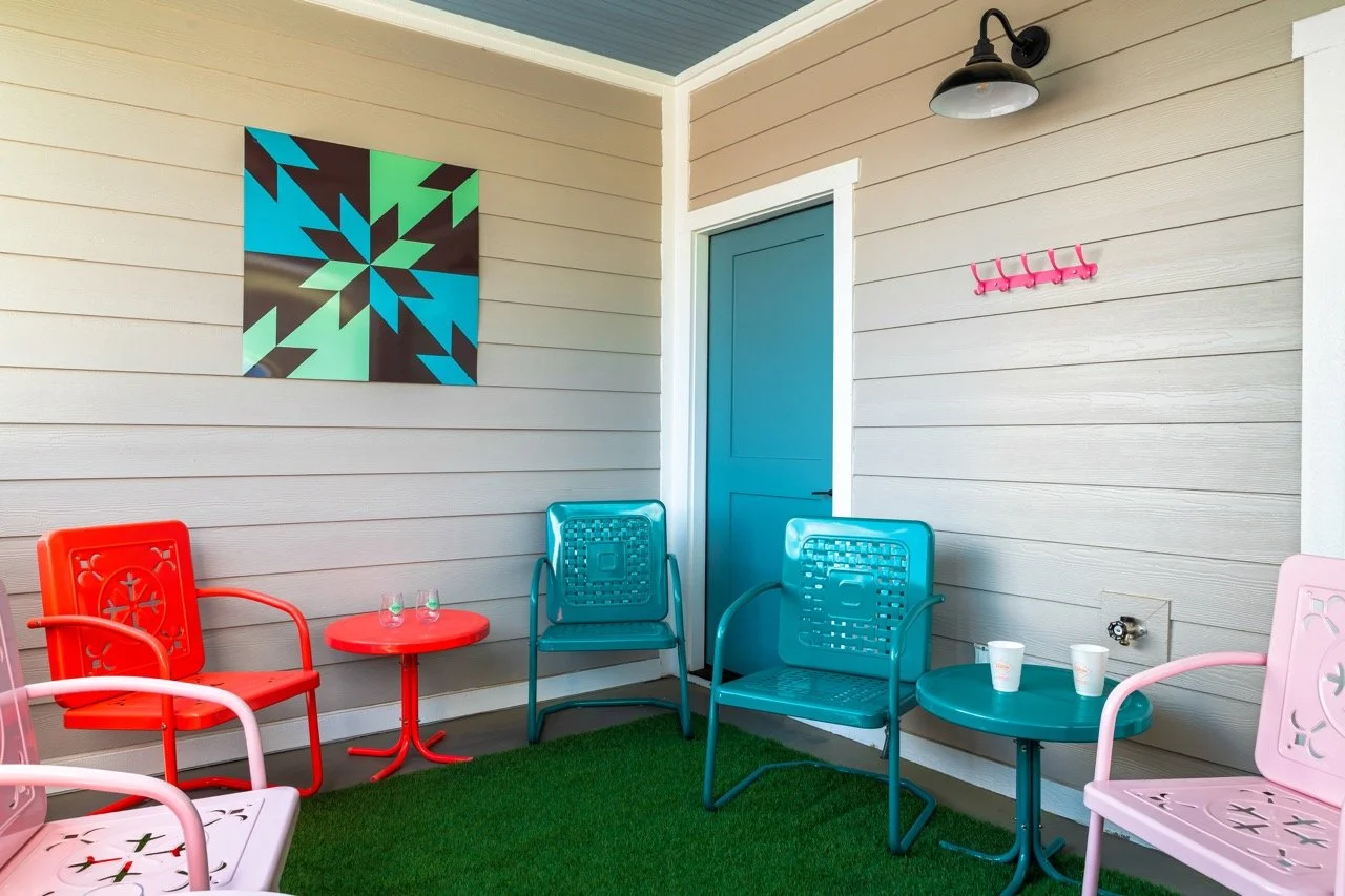 Colorful outdoor patio with plastic chairs in red, pink, teal, and blue around small matching tables, on artificial grass, with a modern wall art, a coat hook, and a wall light.