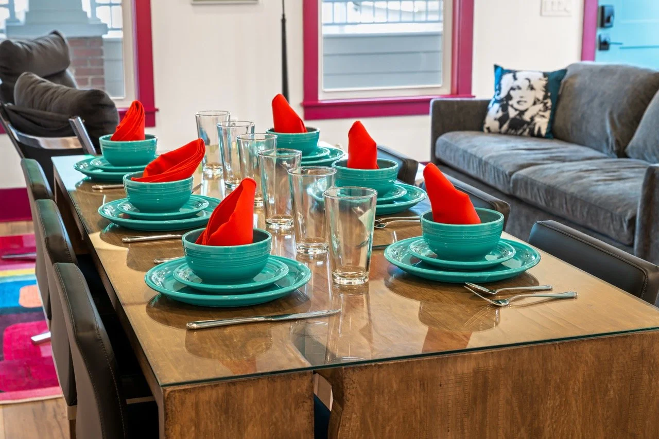 A dining table set for eight with turquoise plates, bowls, red cloth napkins, and clear glasses, in a brightly lit room with pink window trim and a gray couch in the background.