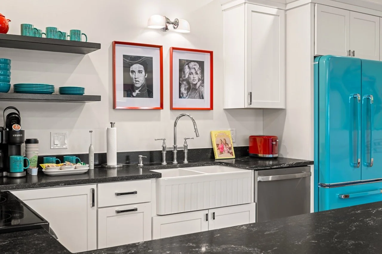 Kitchen with white cabinets, black countertops, a white farmhouse sink, turquoise refrigerator, red toaster, and framed black-and-white portraits on the wall.
