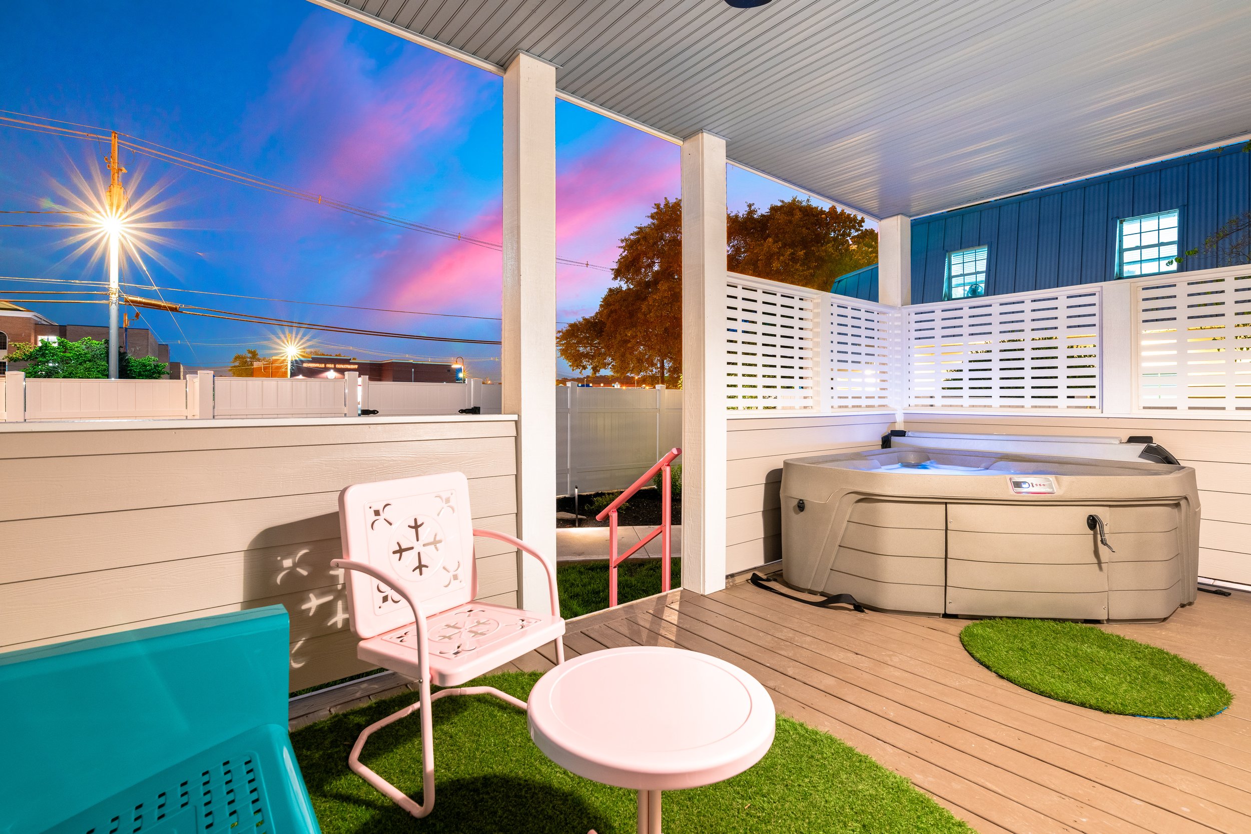 A porch with a hot tub, pink chair, and teal container, with a white fence and a view of trees and lights outside during sunset.