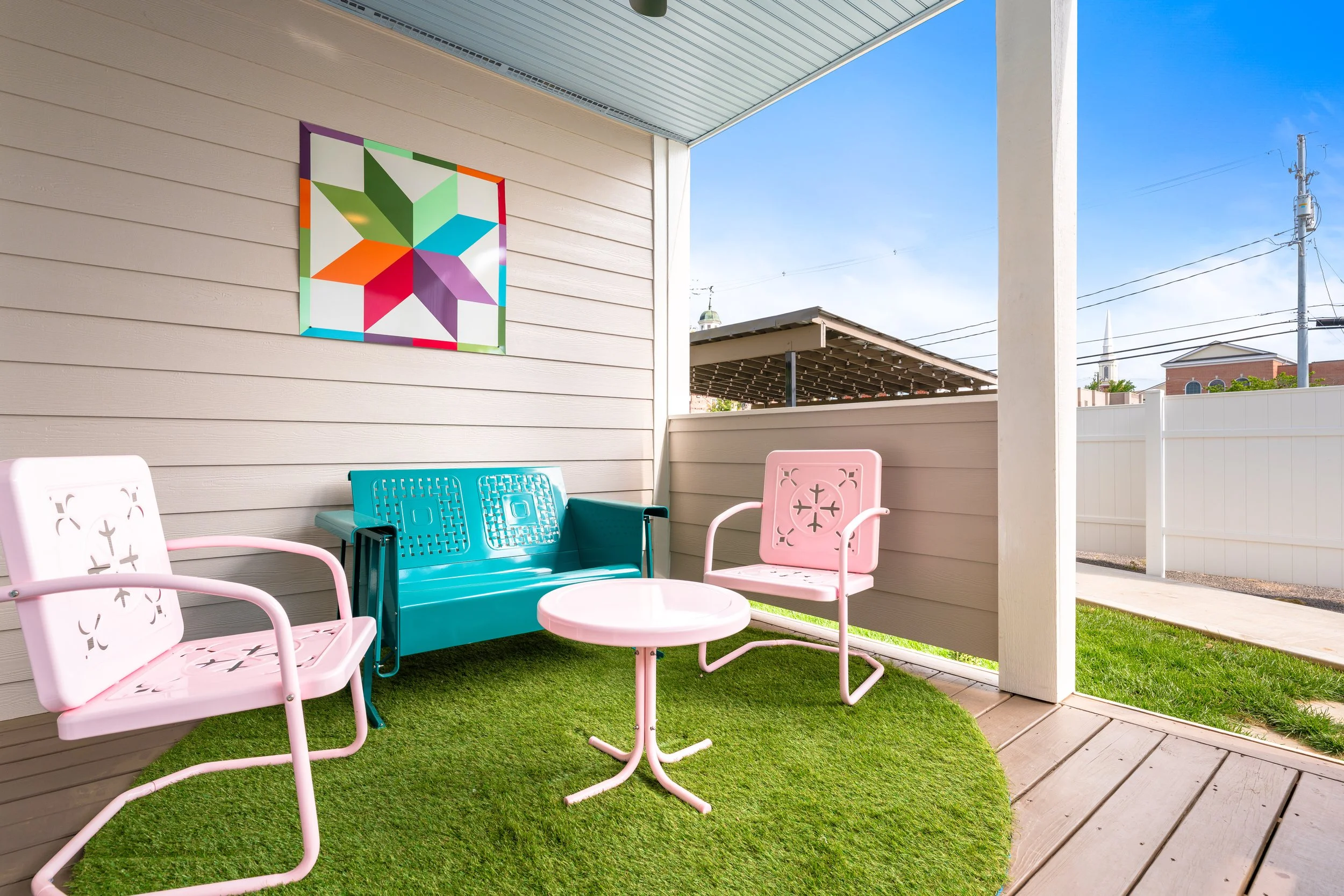 Outdoor patio area with colorful chairs, small pink table, artificial grass, beige wooden wall with a colorful geometric art piece, white fence, and a rooftop with solar panels. In the background, there are utility poles and a church steeple.