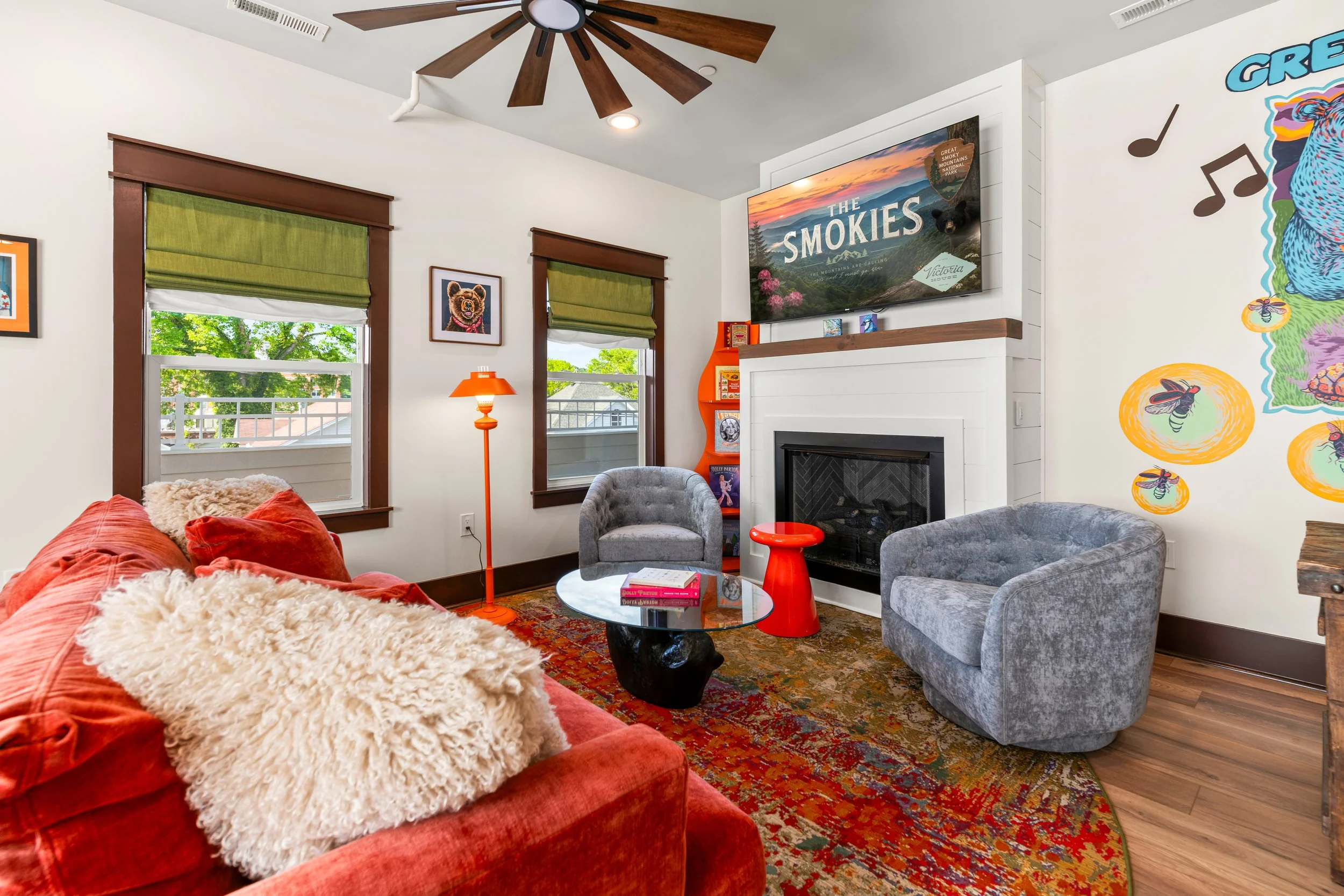 Living room with red sofa covered in beige and white throw blankets, green window shades, framed artwork of a bear on the wall, two gray armchairs, a round glass coffee table with books, a colorful rug, a fireplace, a large TV displaying "The Smokies