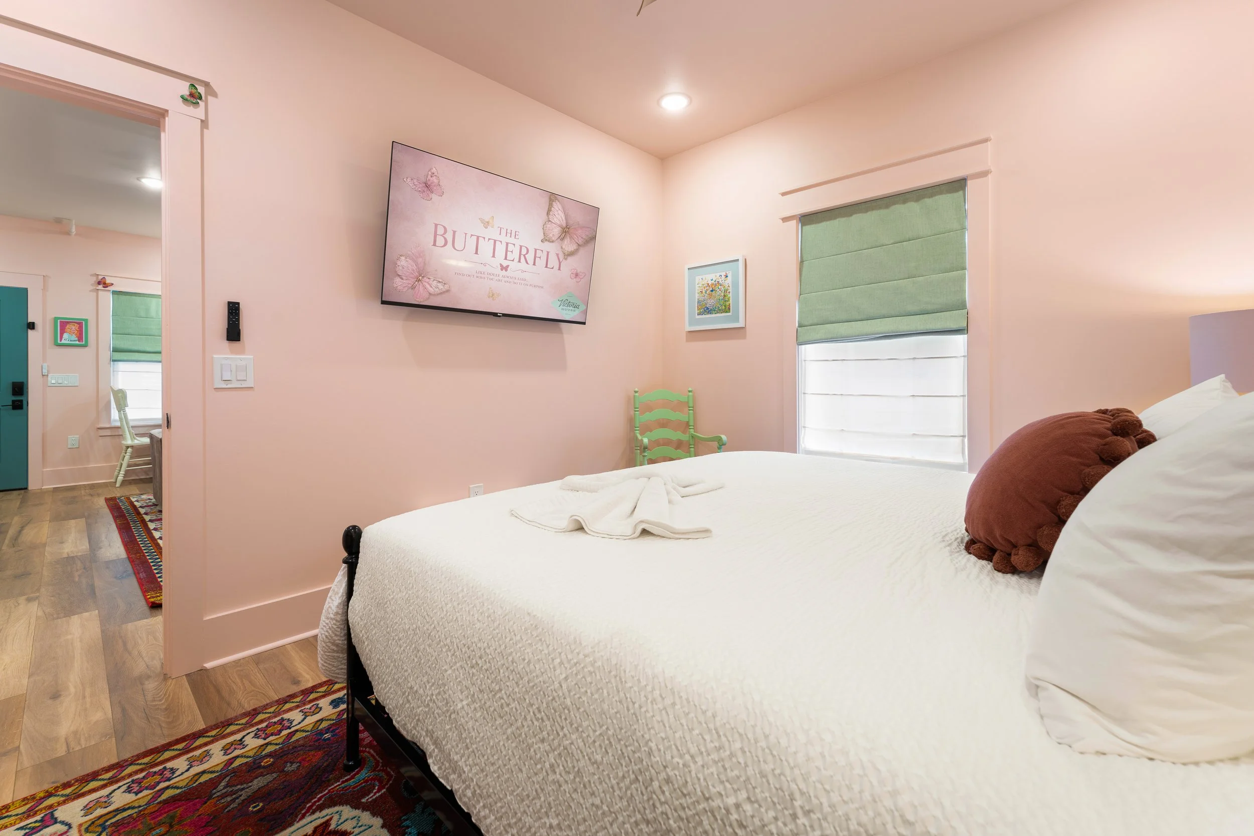 A bedroom with a bed, brown pillows, and quilted white bedding. A green chair is near the wall, which has a framed flower picture. A window with a green Roman shade is on the right side. A flat-screen TV on the pink wall displays a butterfly-themed s