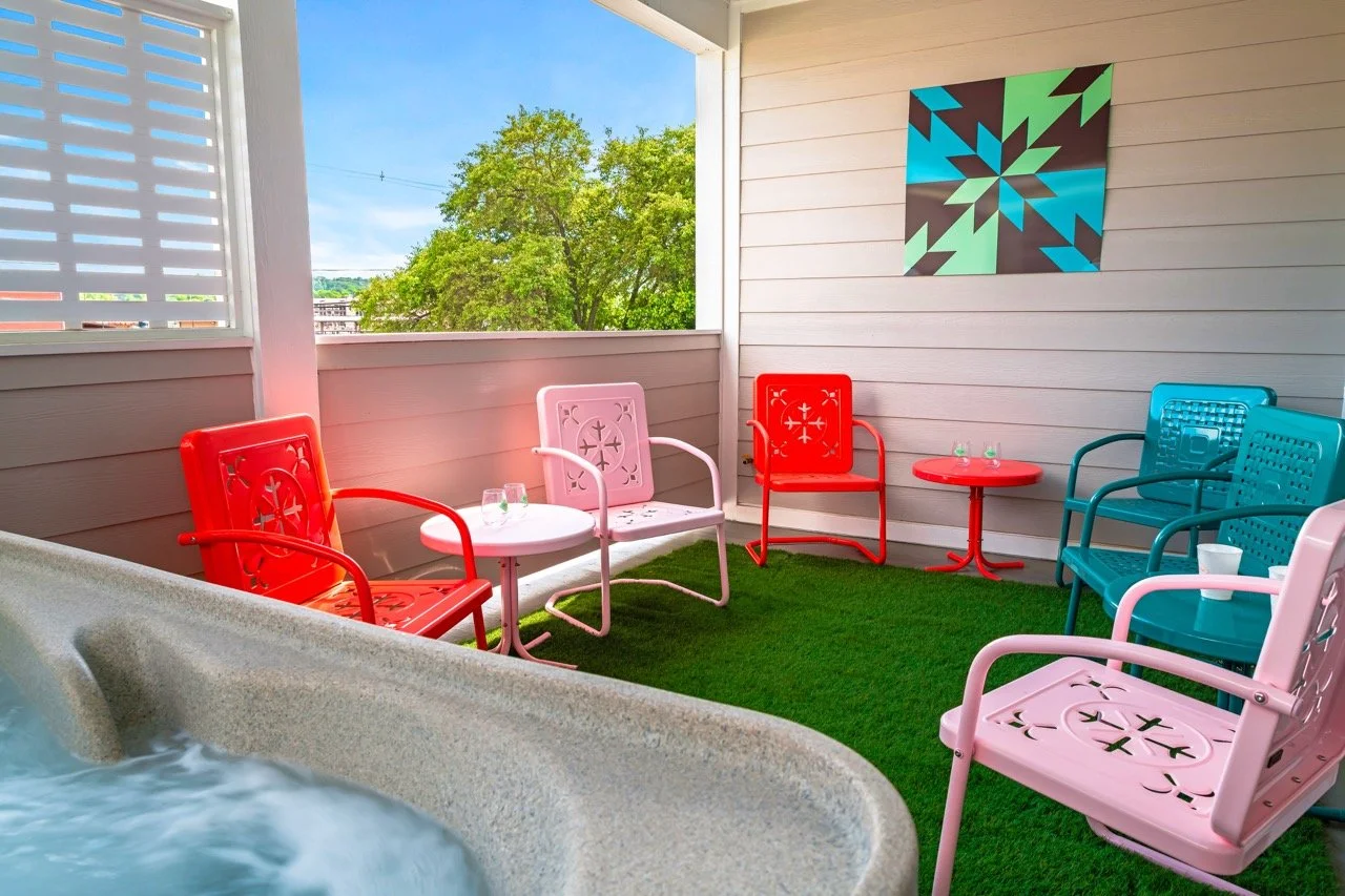 Colorful outdoor patio with pastel pink, red, and blue metal chairs and tables on a green artificial grass area, with a hot tub in foreground, against a background of green trees and a partly cloudy sky.