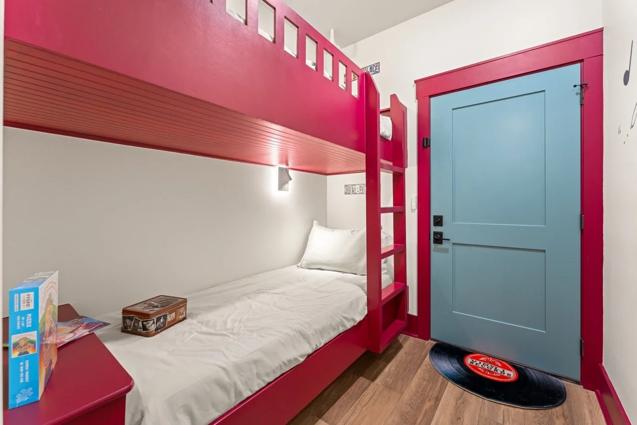 A bedroom with a bunk bed featuring a pink frame, white bedding, and a blue door with pink trim. There is a small ladder attached to access the upper bunk, and toys and a box are on the bed and nightstand. A vinyl record-shaped rug is on the floor in