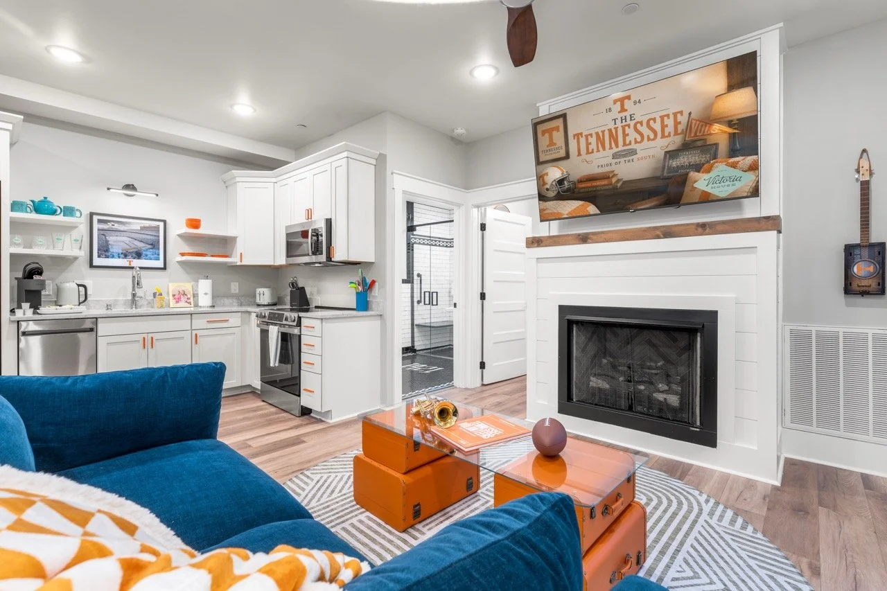 Living room with a white kitchen, fireplace, flat-screen TV, blue sofa, orange trunk coffee table, and Tennessee Volunteer memorabilia.
