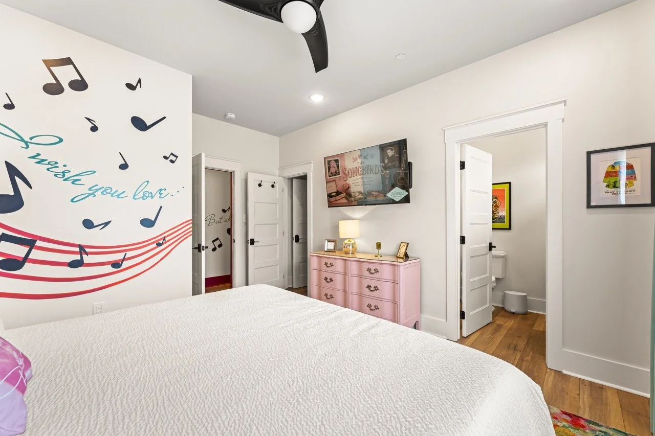 Bedroom with white walls, a large bed with white bedding, a pink dresser, wall art, a mounted TV, and an en-suite bathroom with a toilet.