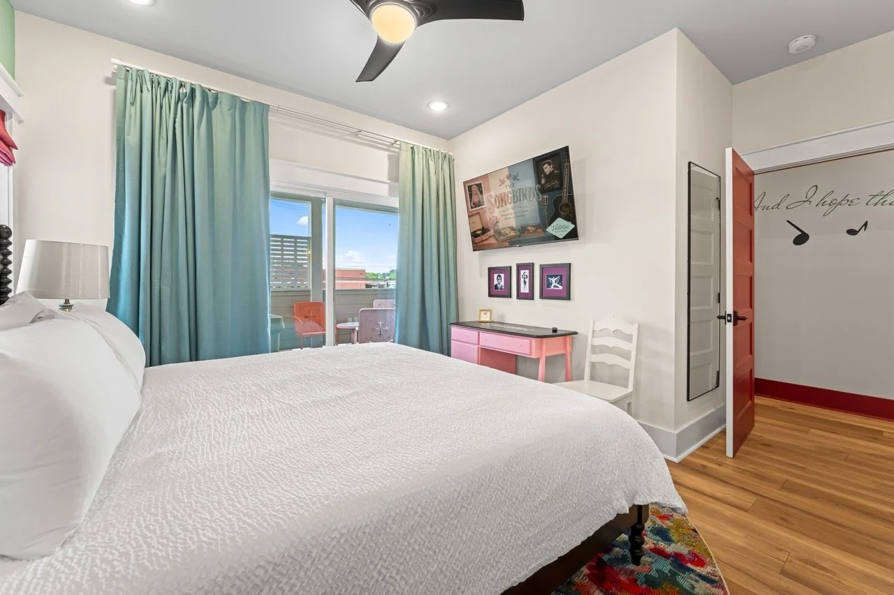 A bedroom with a white bed, colorful curtains, a ceiling fan, a small desk with framed pictures, a white chair, and a sliding glass door leading to a balcony with outdoor seating.