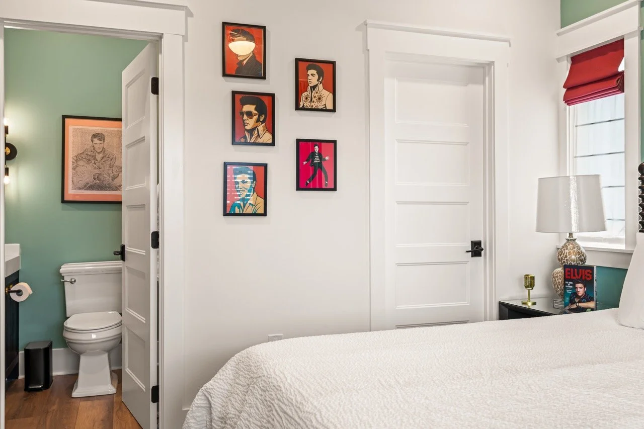 A bedroom with artwork of Elvis Presley on the wall, a bed with a white quilt, and a nightstand with a lamp, a book titled 'Elvis,' and a small golden object. An adjacent bathroom with a toilet and framed art is visible.
