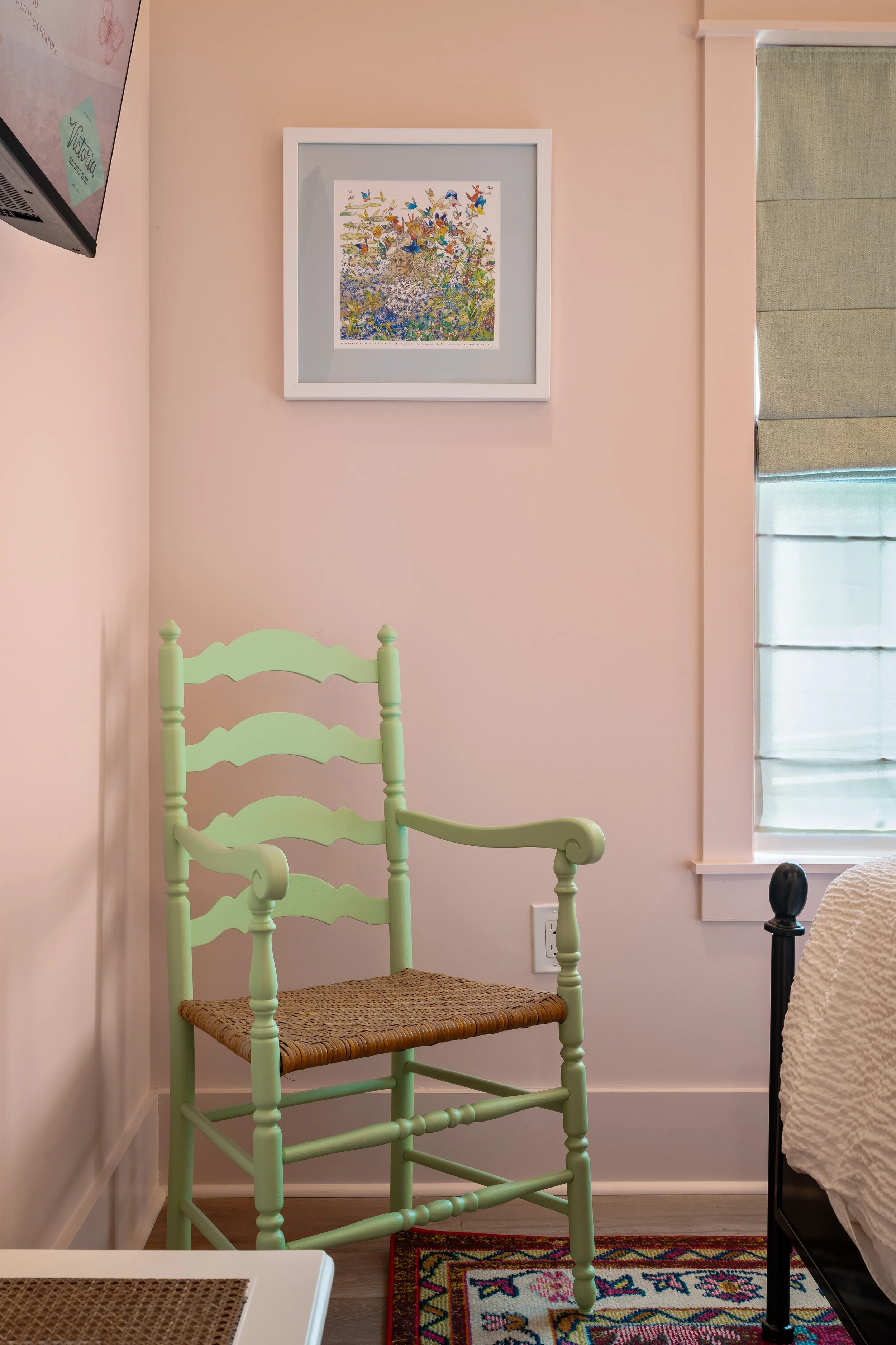 A light green wooden chair with woven seat next to a bed with white bedding and a black post, placed against a light pink wall with a framed colorful artwork of butterflies and a woman, and a window with beige blinds and a colorful rug on the floor.
