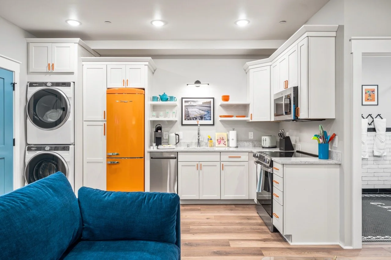 Kitchen with white cabinets, orange refrigerator, stainless steel appliances, open shelving, and a blue couch in the foreground.