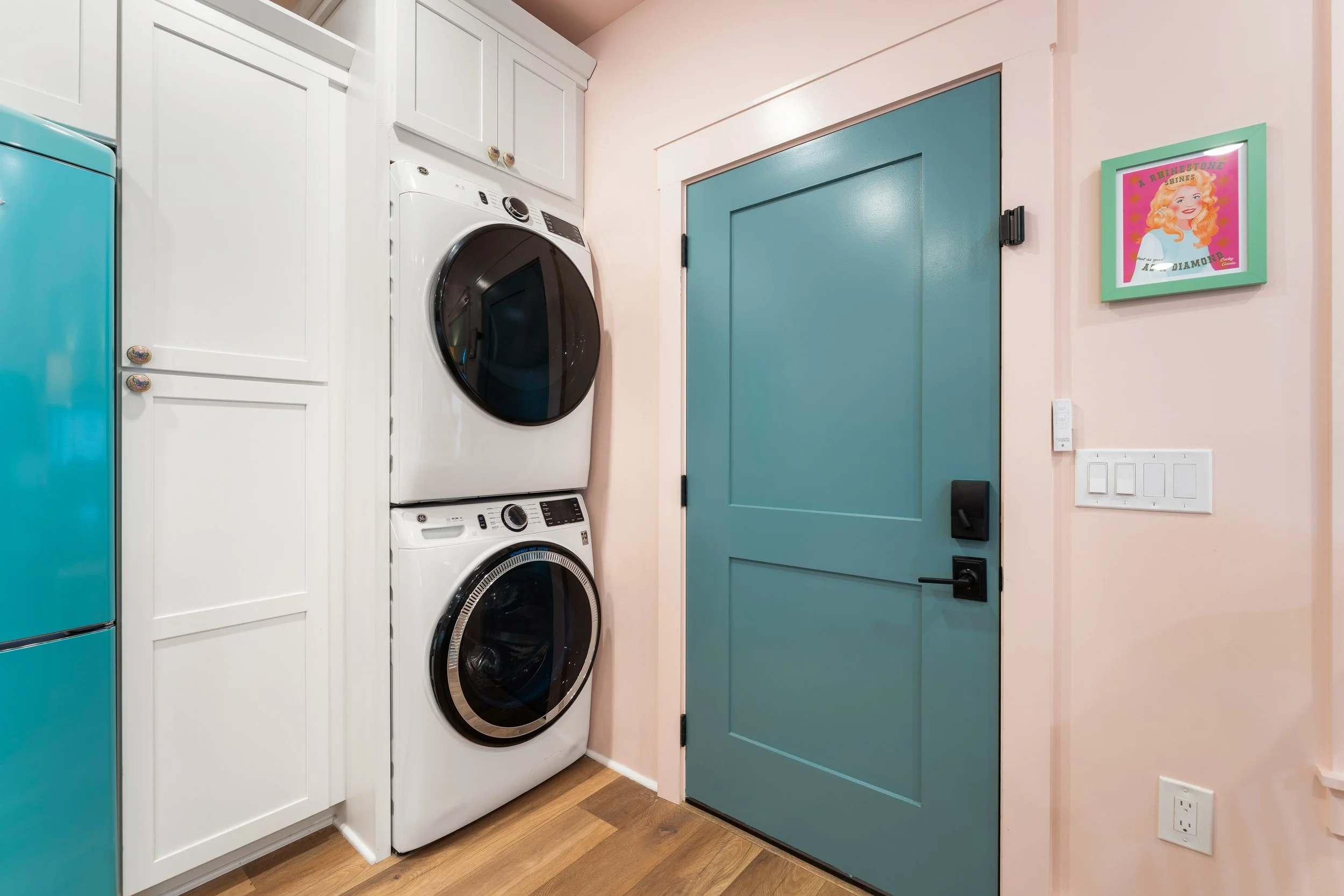 A laundry area with a teal refrigerator, white cabinets, a stacked front-loading washer and dryer, a teal sliding door, and a framed picture of a woman with red hair on the wall.
