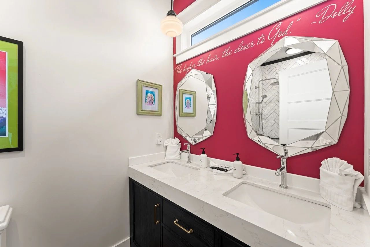 Double vanity bathroom with two geometric mirrors, pink accent wall with quote, white countertop, and framed artwork