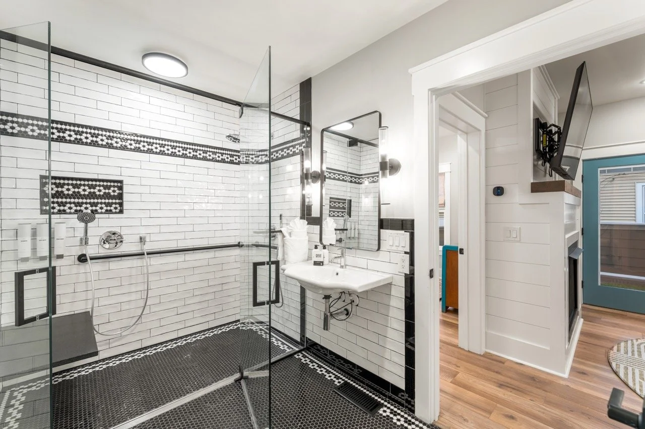 Bathroom with walk-in glass shower enclosure, white subway tile walls with black accents, a small white sink, and a wooden floor.