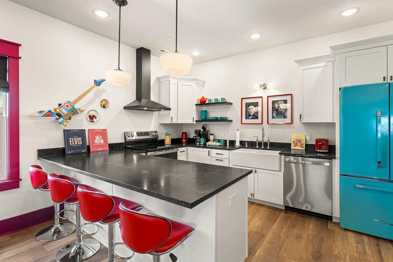 A bright, modern kitchen with white cabinets, black countertops, and colorful retro appliances. Features red bar stools along a black-topped island, vintage decor including a guitar wall art, Elvis and Dolly Parton books, and framed black-and-white p