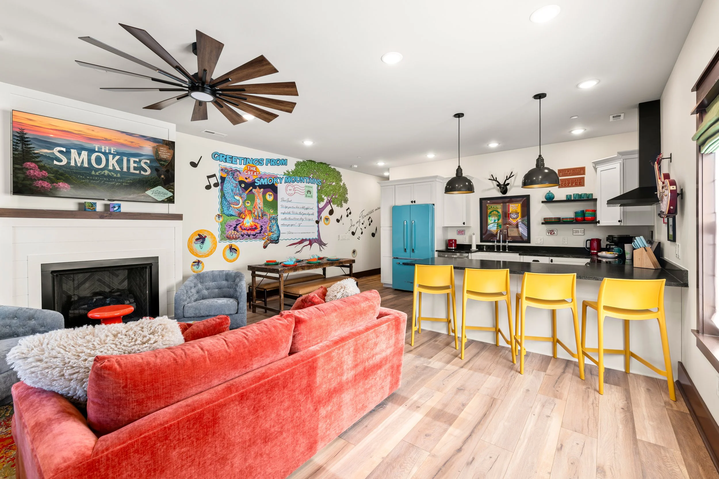 Interior of a modern, colorful living room and kitchen with a red couch, gray armchair, and yellow bar stools, decorated with musical and mountain-themed posters.