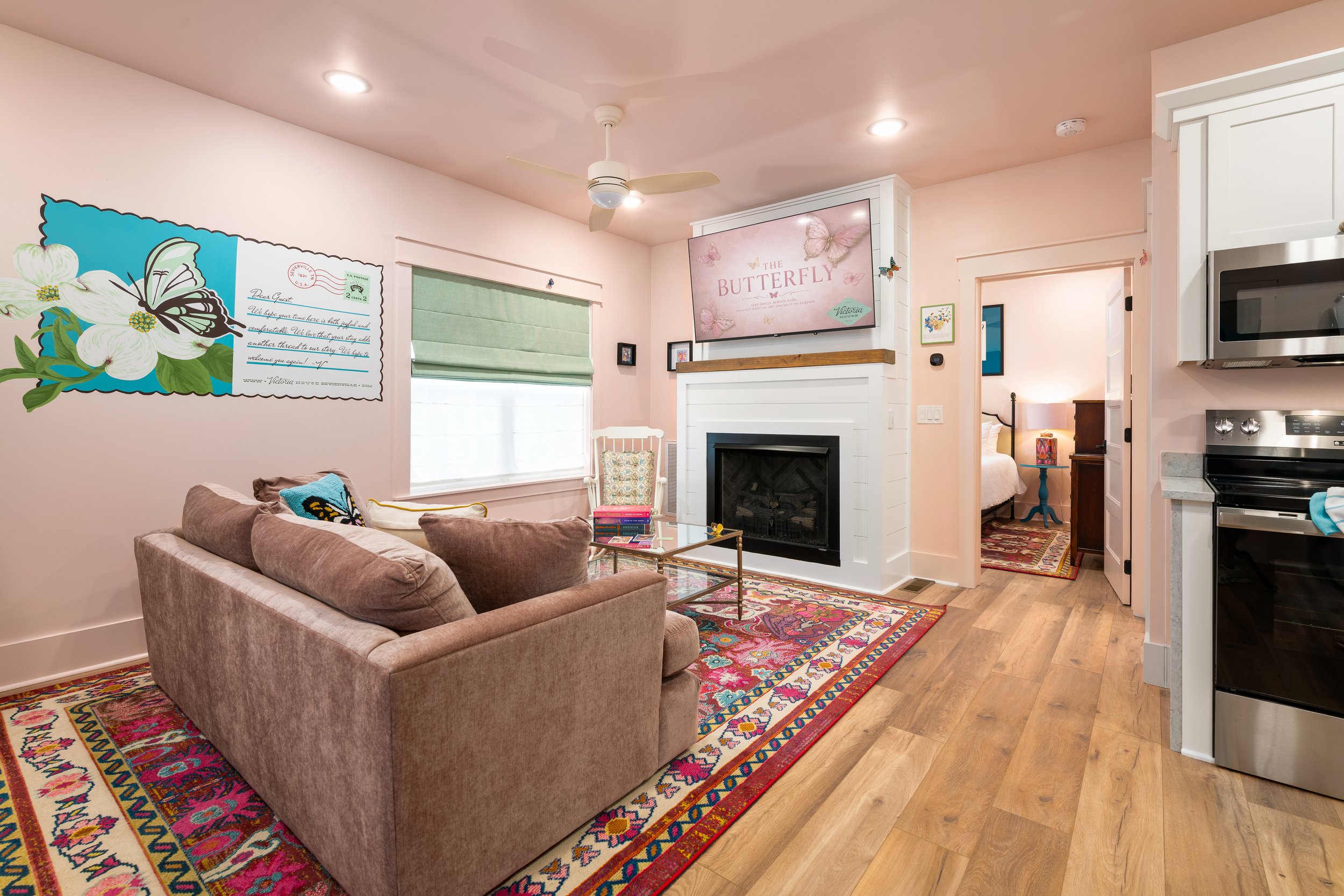 Living room with a brown couch, colorful patterned rug, fireplace, TV, ceiling fan, and a bedroom visible through an open doorway.
