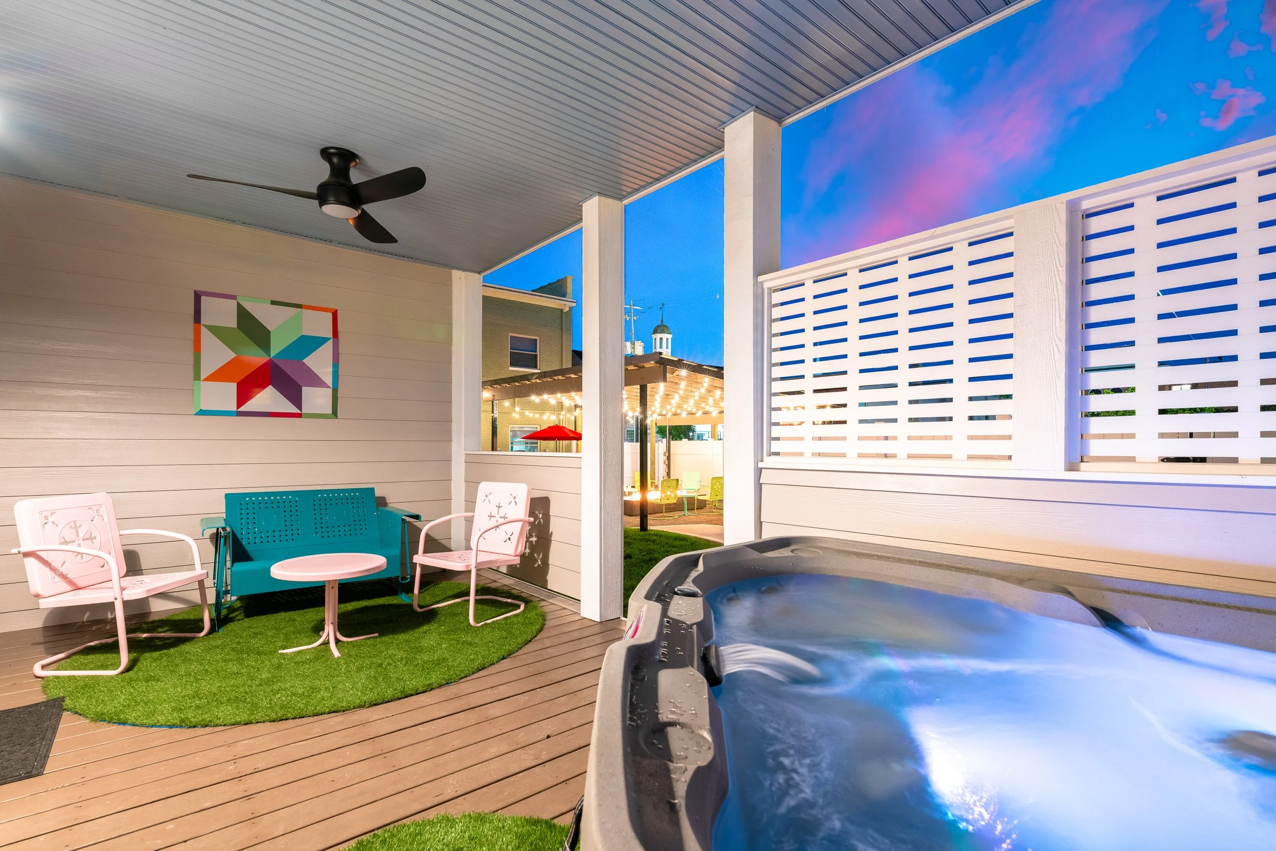 A covered outdoor patio area with a ceiling fan, colorful wall art, pink and turquoise chairs with a small table, a green artificial grass rug, a hot tub, and a privacy fence. In the background, there are string lights and umbrellas, with a clear eve
