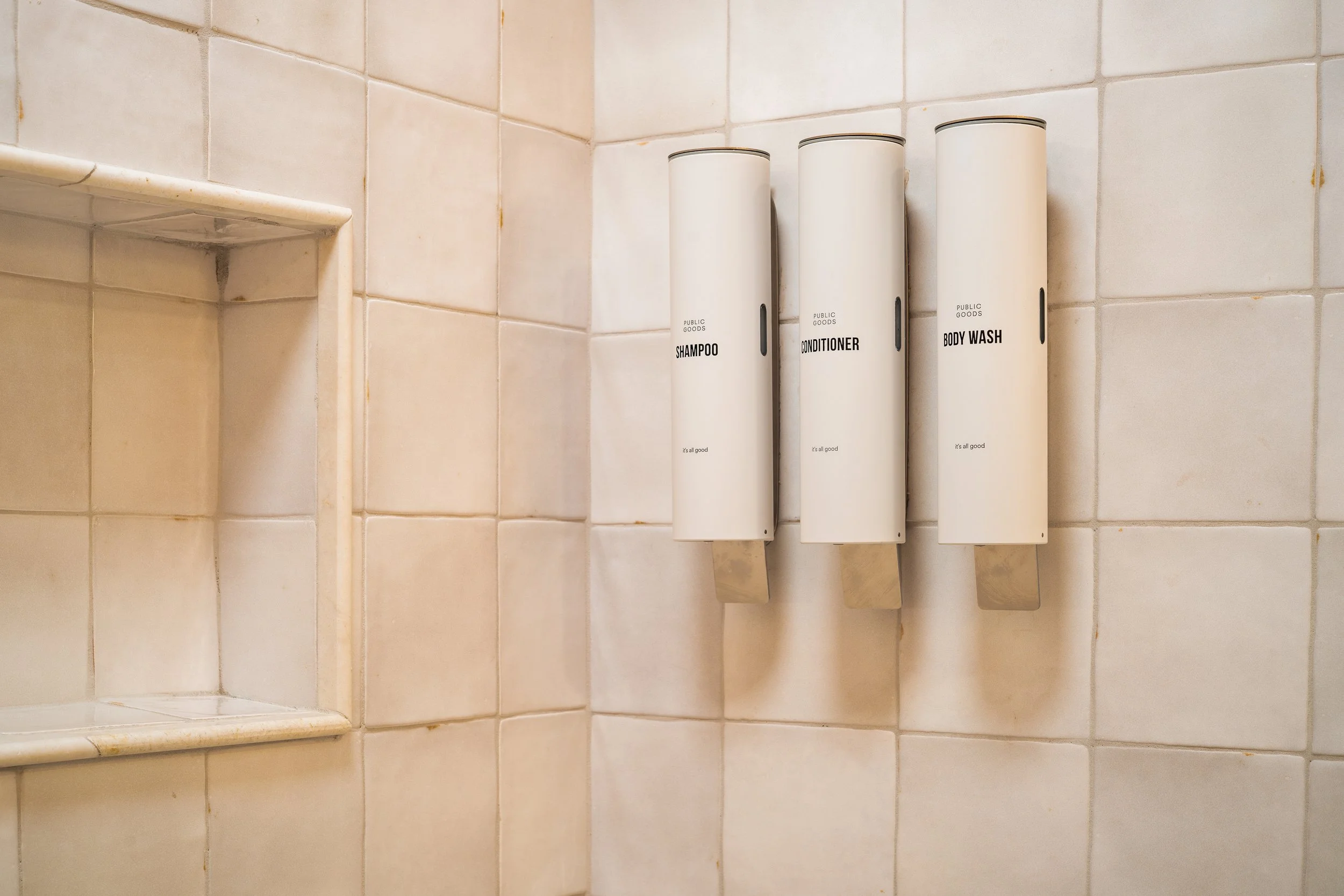 Three white bottles labeled 'Shampoo', 'Conditioner', and 'Body Wash' are placed vertically against a beige tiled wall in a shower area.