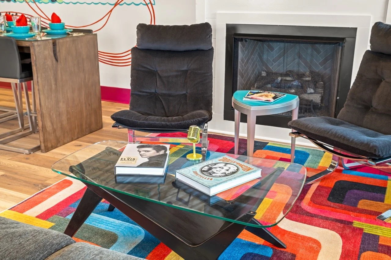 Living room with black chairs, a glass coffee table with Elvis Presley and Marilyn Monroe books, a fireplace, colorful geometric rug, and a partially visible wooden dining table set with plates and glasses.