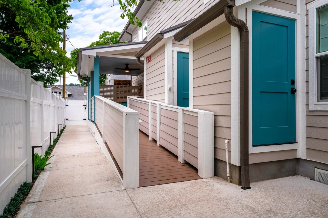 A new residential apartment building with beige siding and teal doors, featuring a concrete walkway, white fencing, and a small ramp for wheelchair access, with trees and power lines in the background.