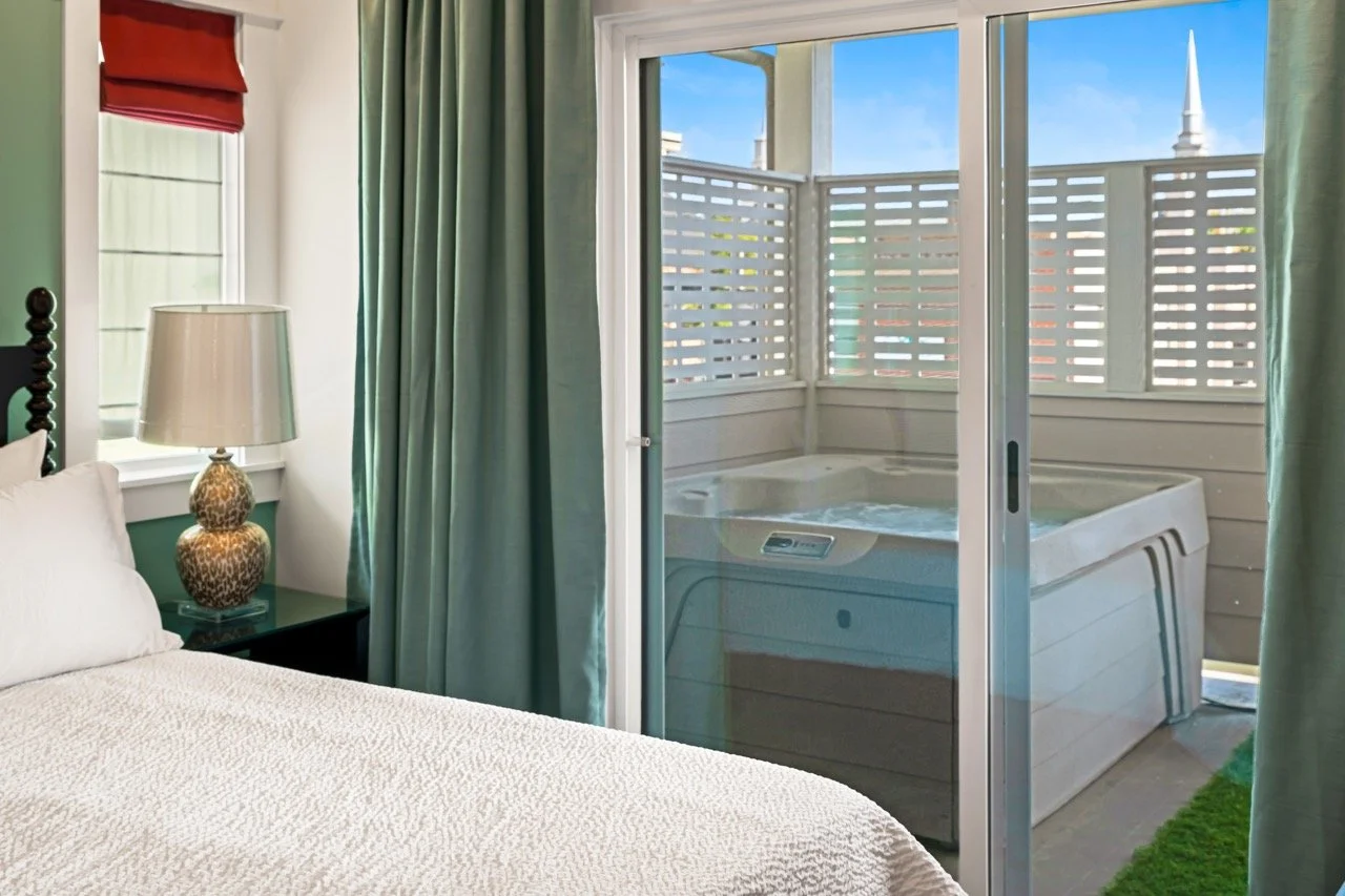 Bedroom with a bed, nightstand with a lamp, window with red curtain, sliding glass door leading to a balcony hot tub with privacy fence, green curtains, blue sky outside.