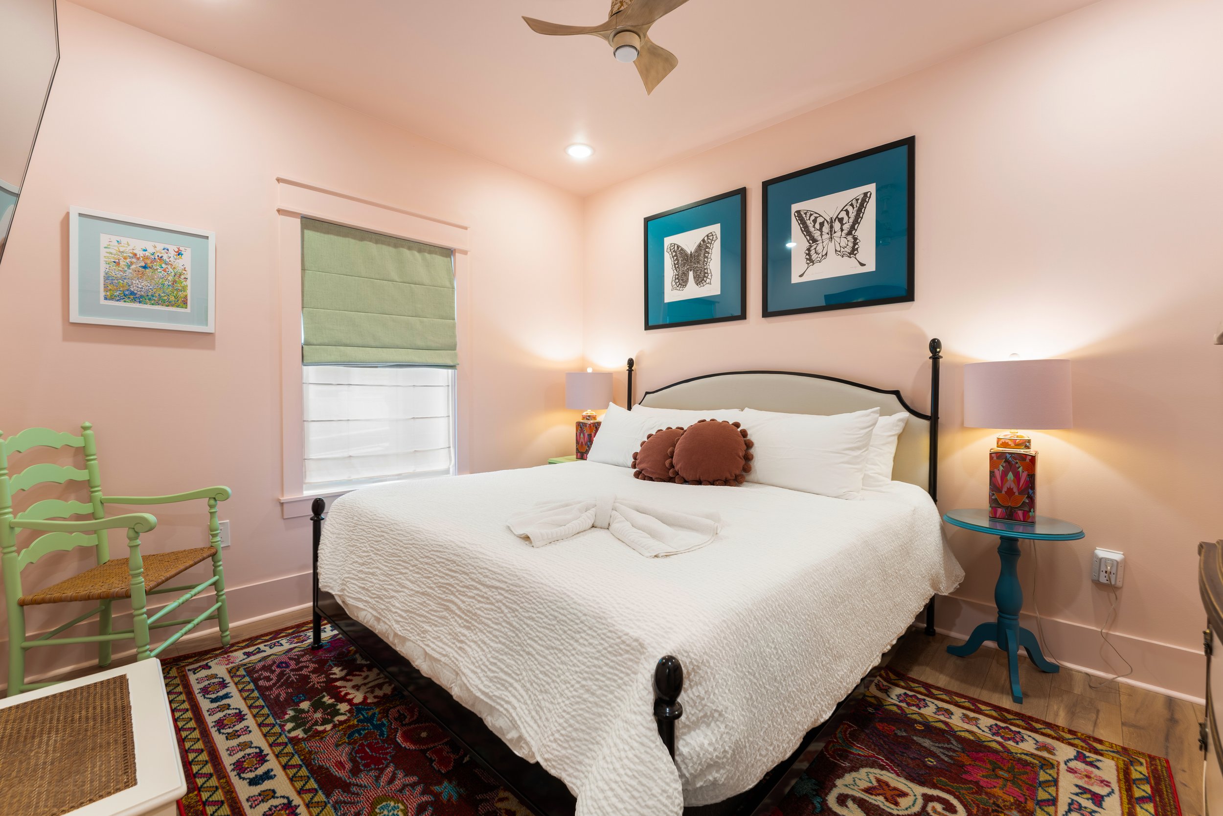 A cozy bedroom with a white bed, brown pillows, a blue side table, and colorful art on the walls. There are two nightstands with lamps, a ceiling fan, and a window with a light green shade.