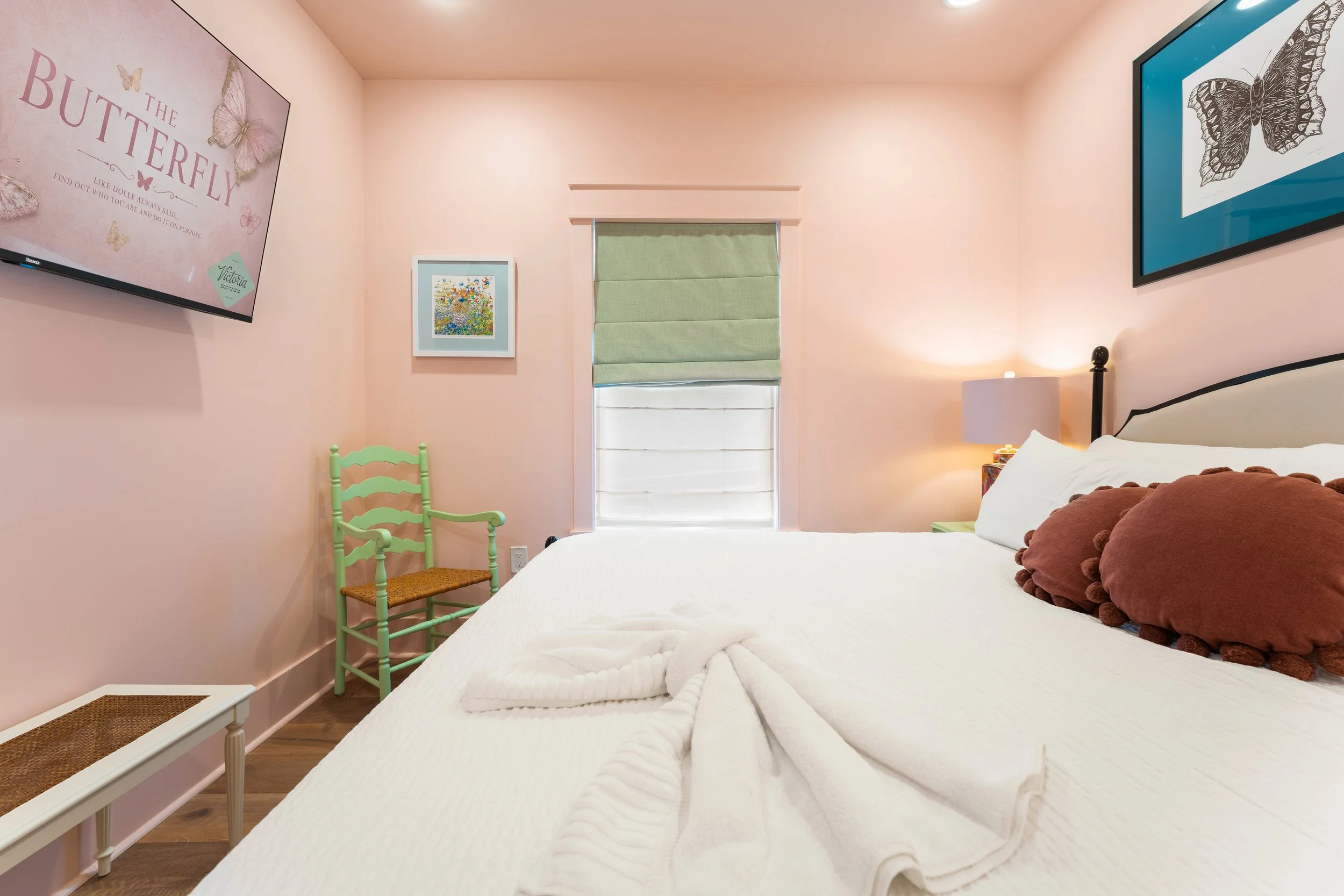 A cozy bedroom with pale pink walls decorated with butterfly artwork and framed pictures. A window with a green Roman shade, a white bed with brown pillows, and a small bedside table with a lamp are visible.