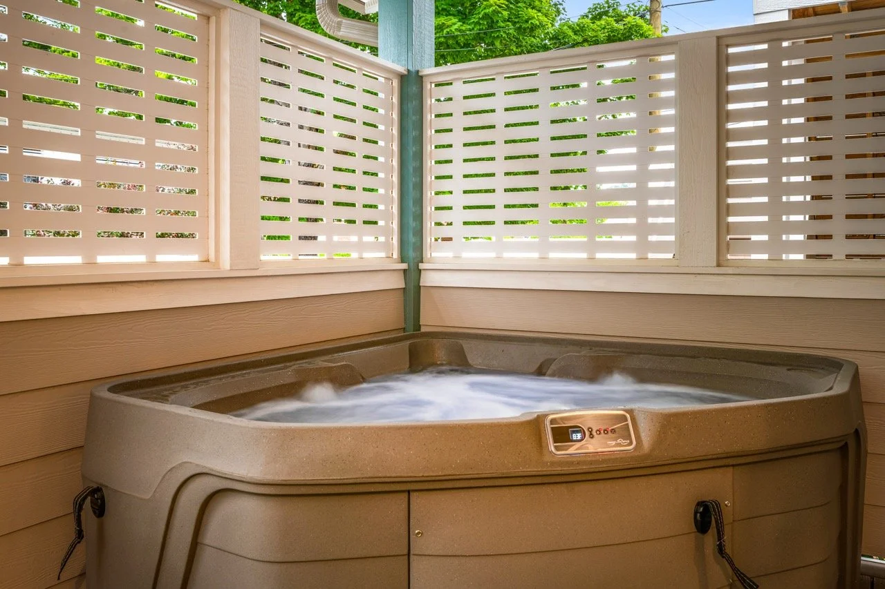 A hot tub on a porch with beige siding, surrounded by white lattice privacy fencing, with green trees visible in the background.