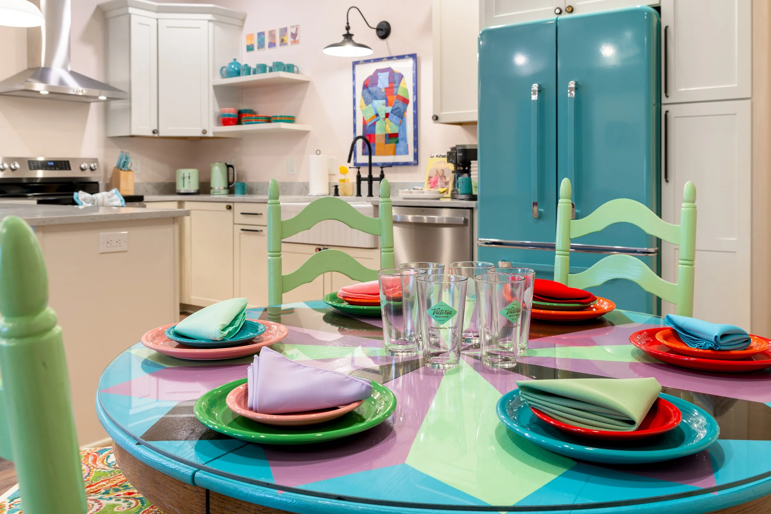 Colorful dining table with place settings, glasses, and napkins in a kitchen with white cabinets, teal fridge, and wall art.