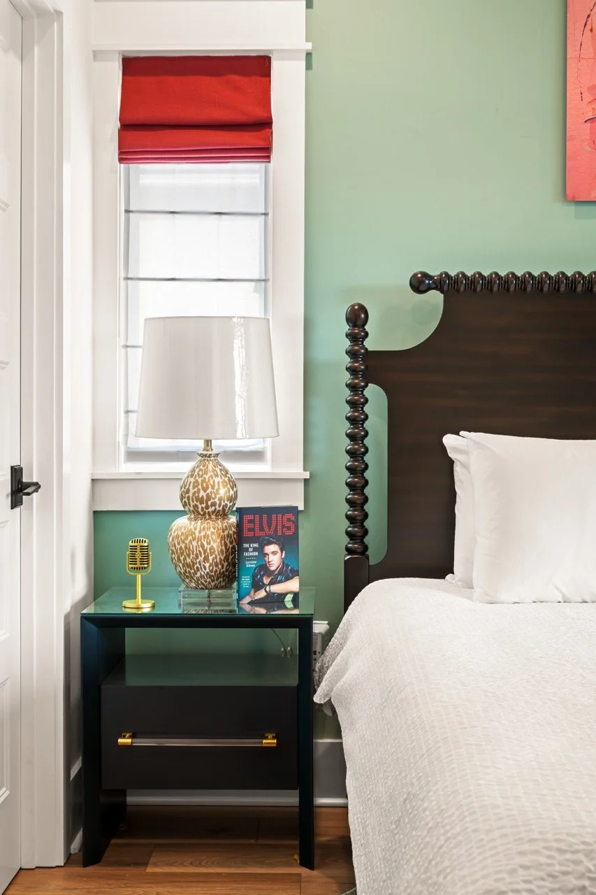 A bedroom corner with a dark wooden bed frame, a white bedding, a black nightstand with a gold handle, a leopard print table lamp, a yellow vintage microphone, a magazine featuring Elvis Presley, a window with white blinds and red Roman shade, and a 